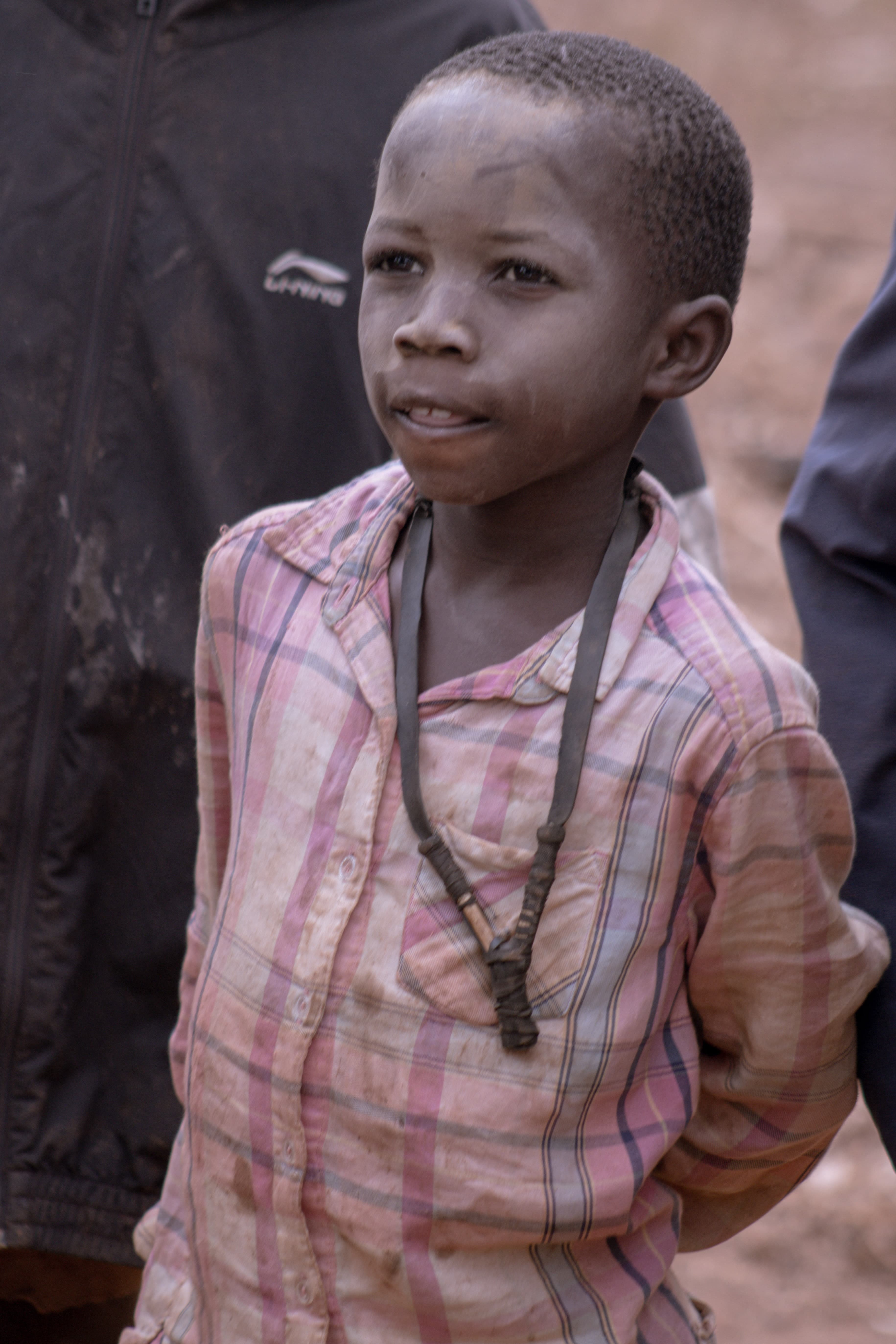 A young girl in a plaid dress wears a beaded necklace and stands on a dirt road, looking off to the side.