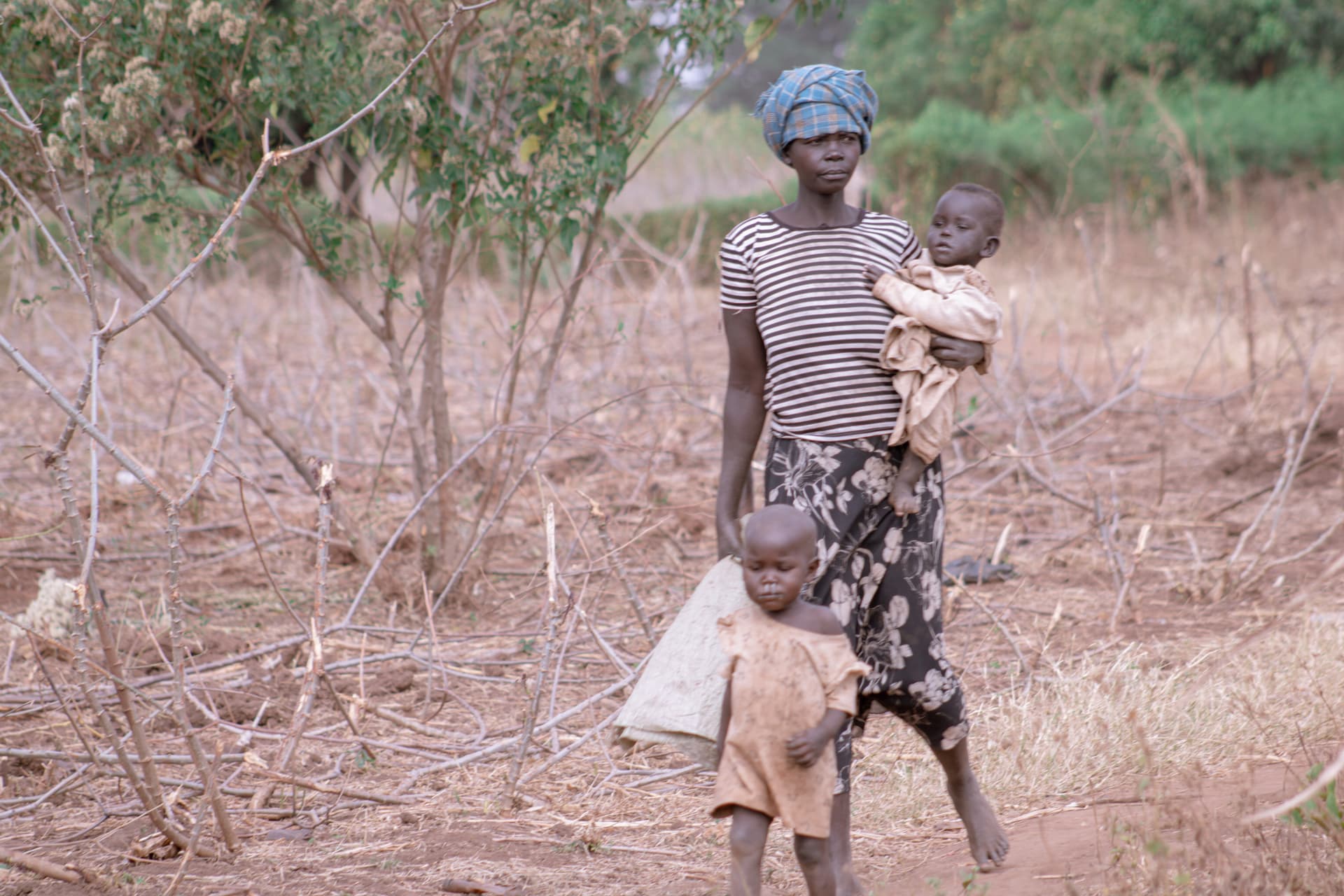 A woman carries a baby and a bag while walking through dry brush.
