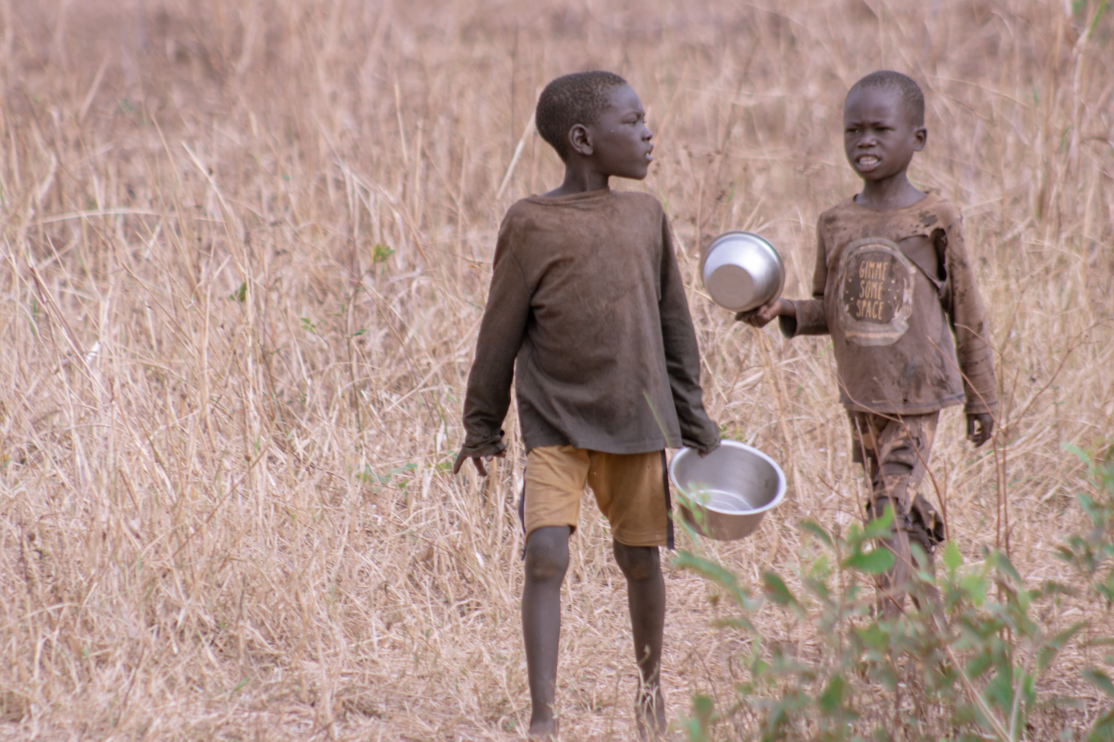 Two boys stand in a field, one holding a metal cup and a bowl.