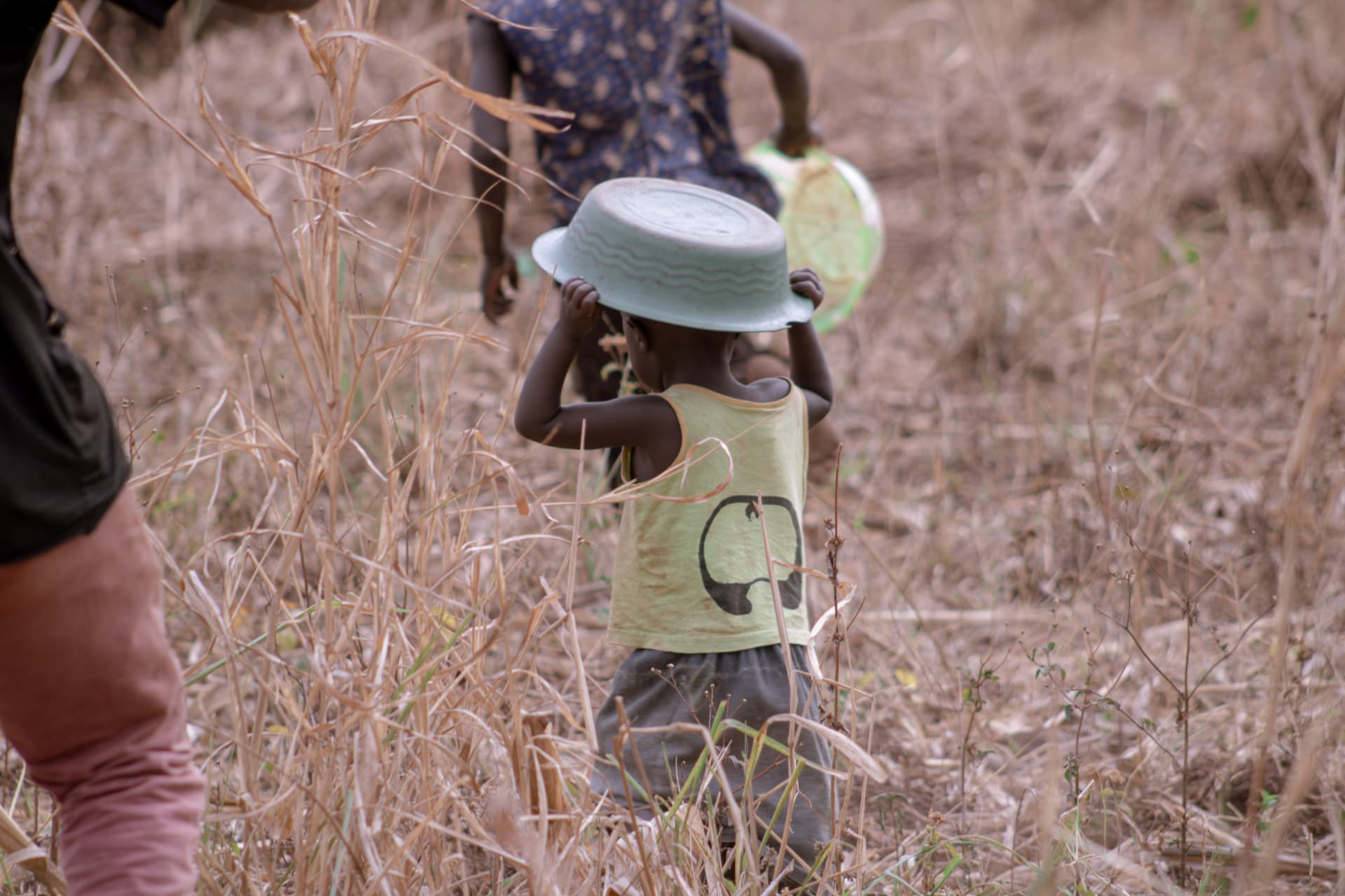 A child walks through tall dry grass balancing a metal bowl on their head.