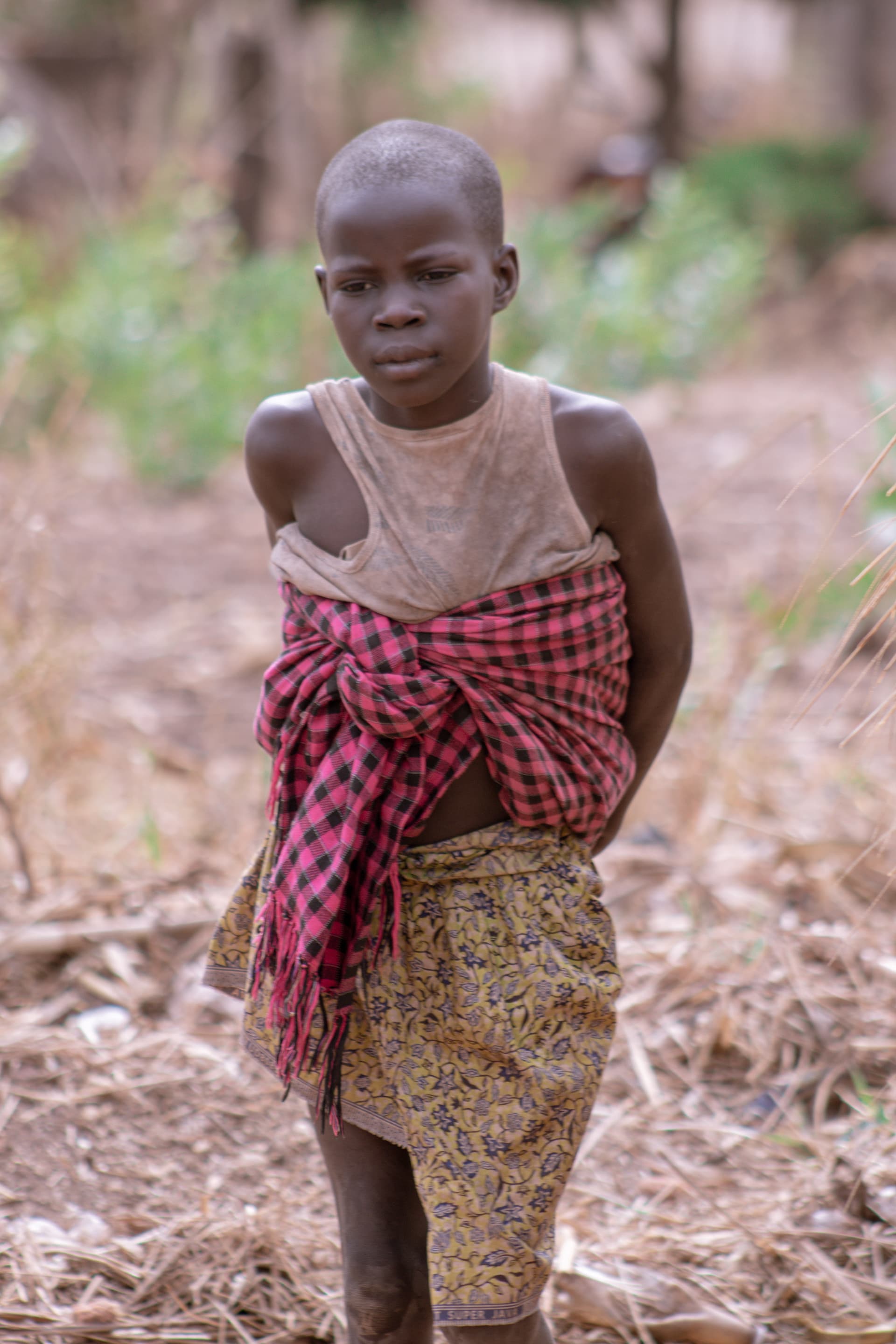 A child stands alone in dry grass wearing a red plaid wrap, in the village of Isunga.
