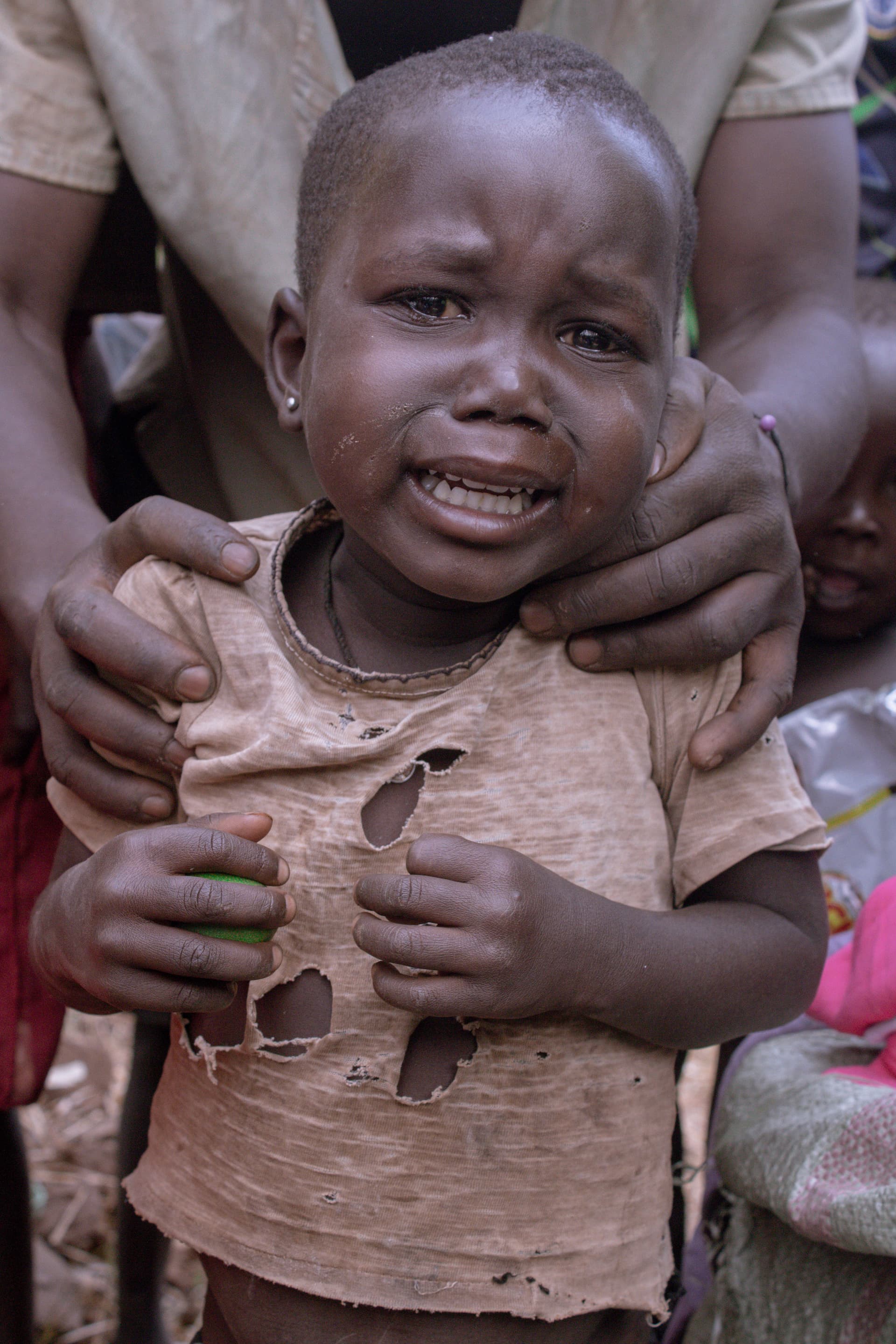Close-up of a crying young child in a torn shirt being held by an adult.