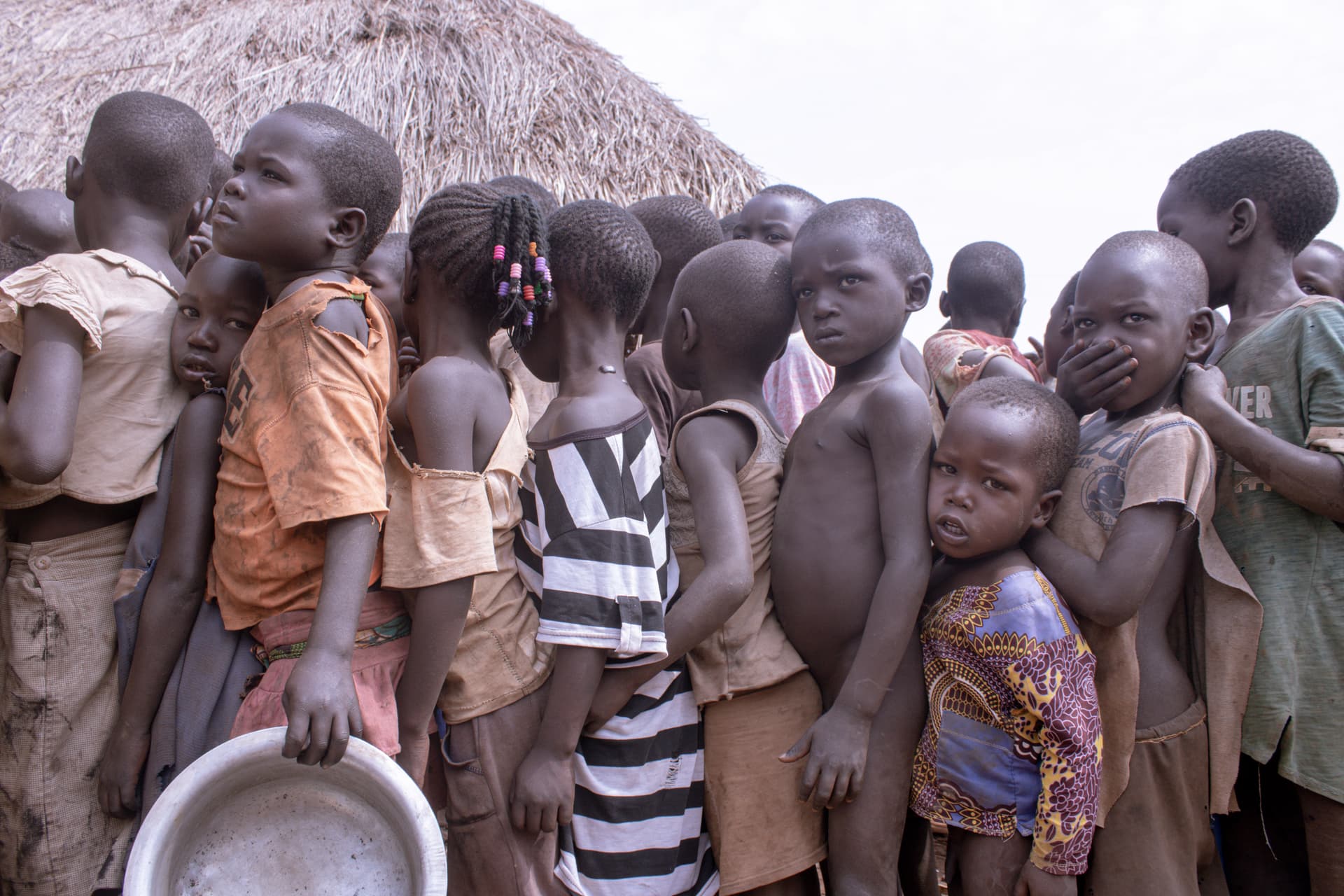 A line of children waits outdoors near a thatched structure, one holding a metal basin.