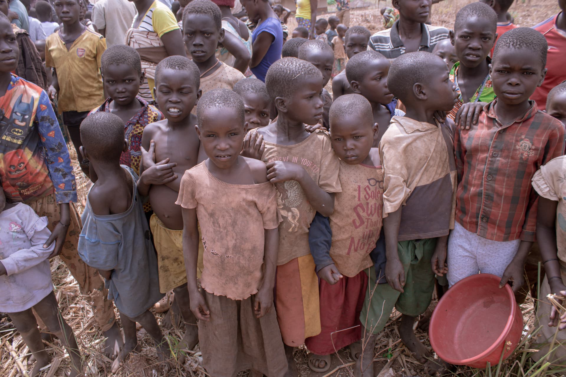 A group of children stand together outdoors, one holding a red plastic basin.