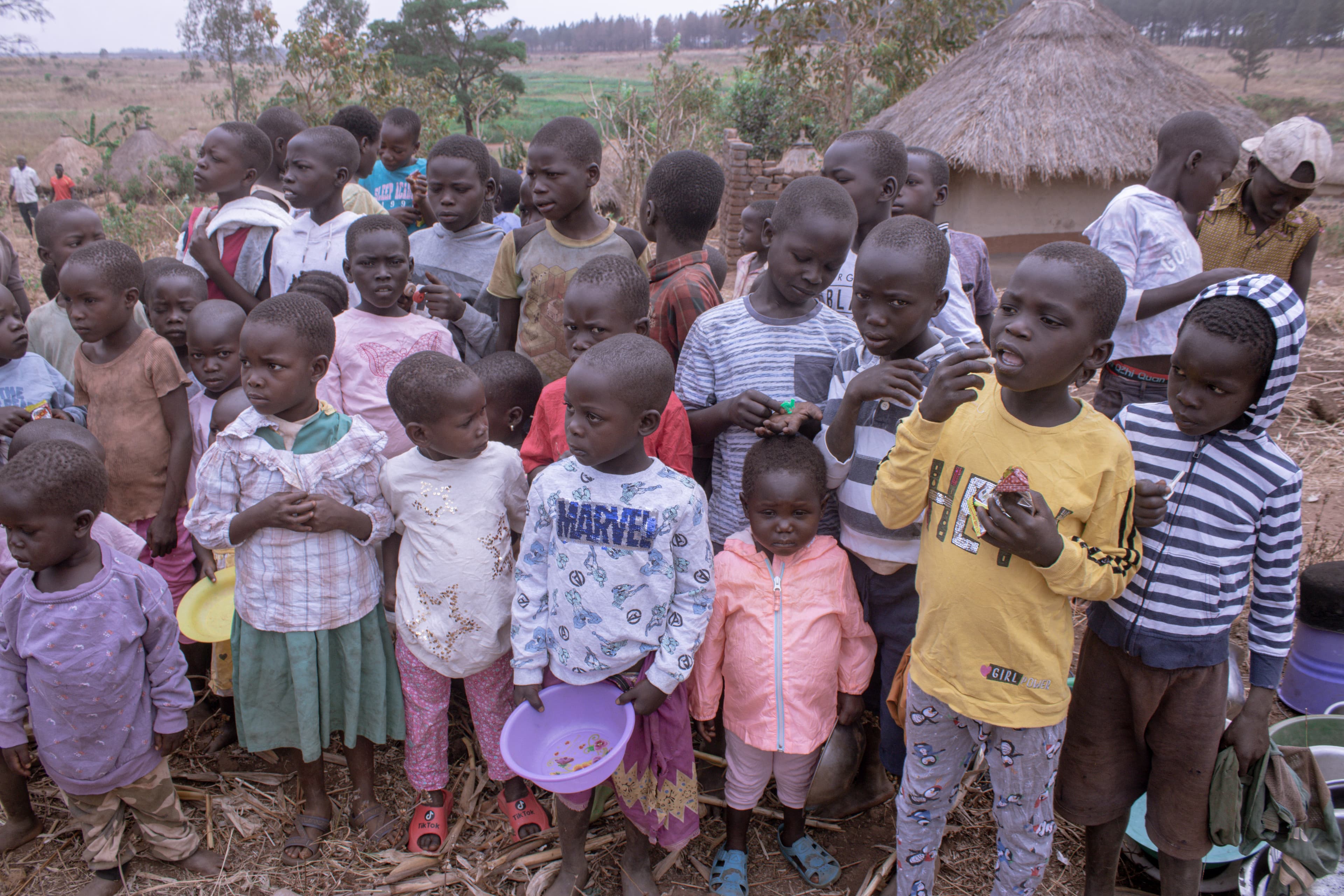 A group of children in mismatched clothing stand together near village huts.