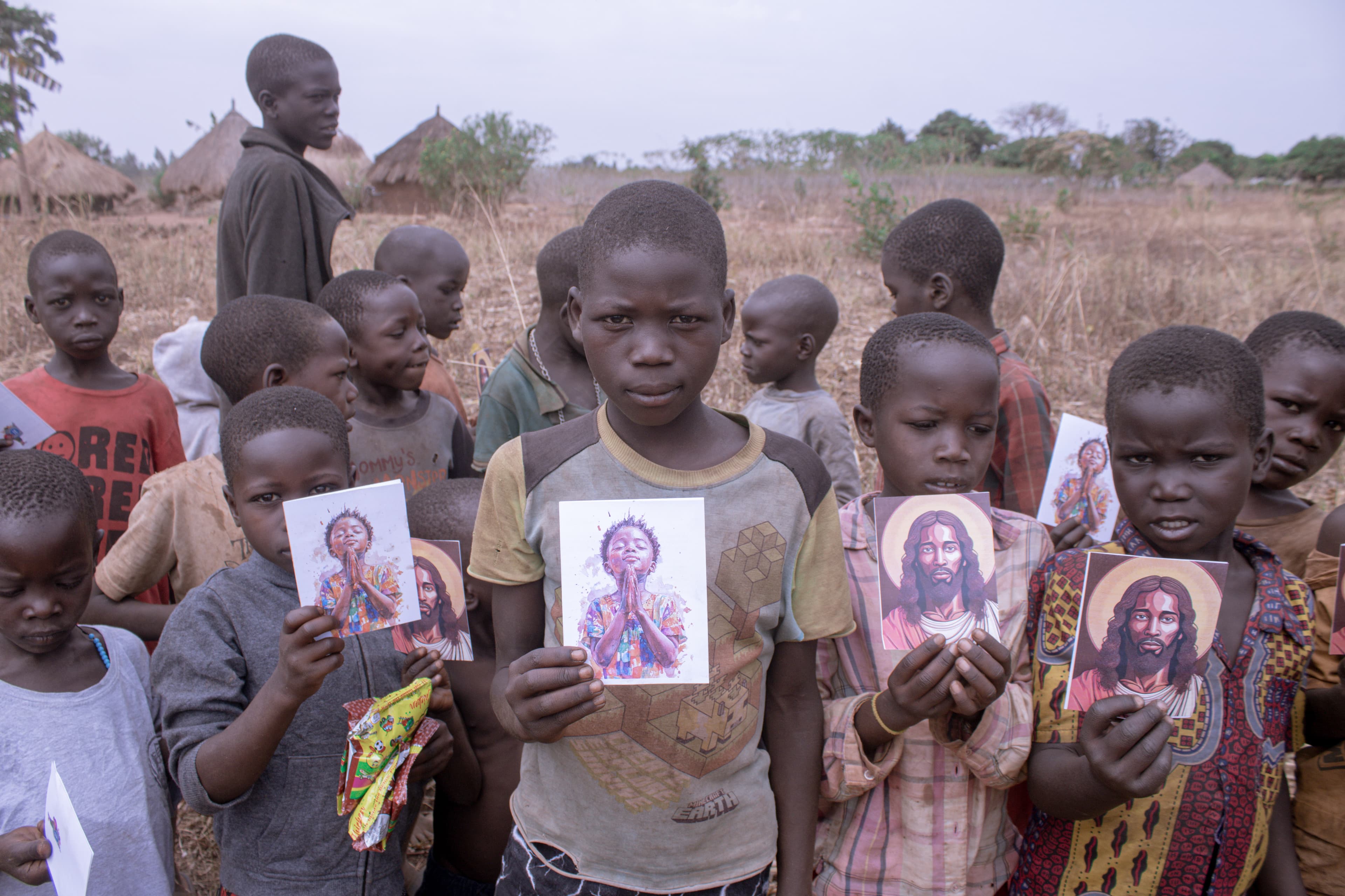 A crowd of children and adults hold up printed devotional cards outdoors near a village.
