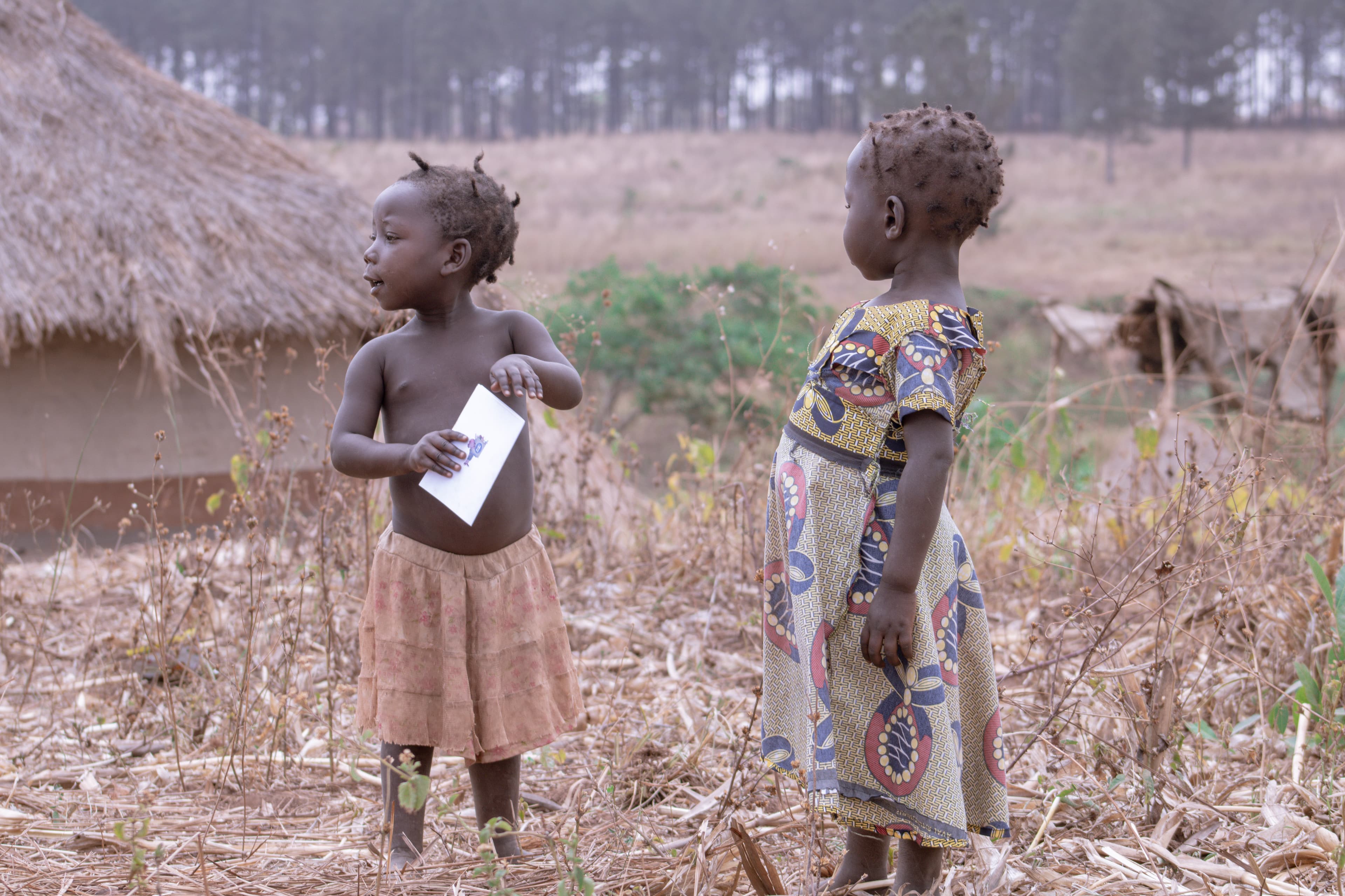 Two young children stand together near huts, one holding a printed card.