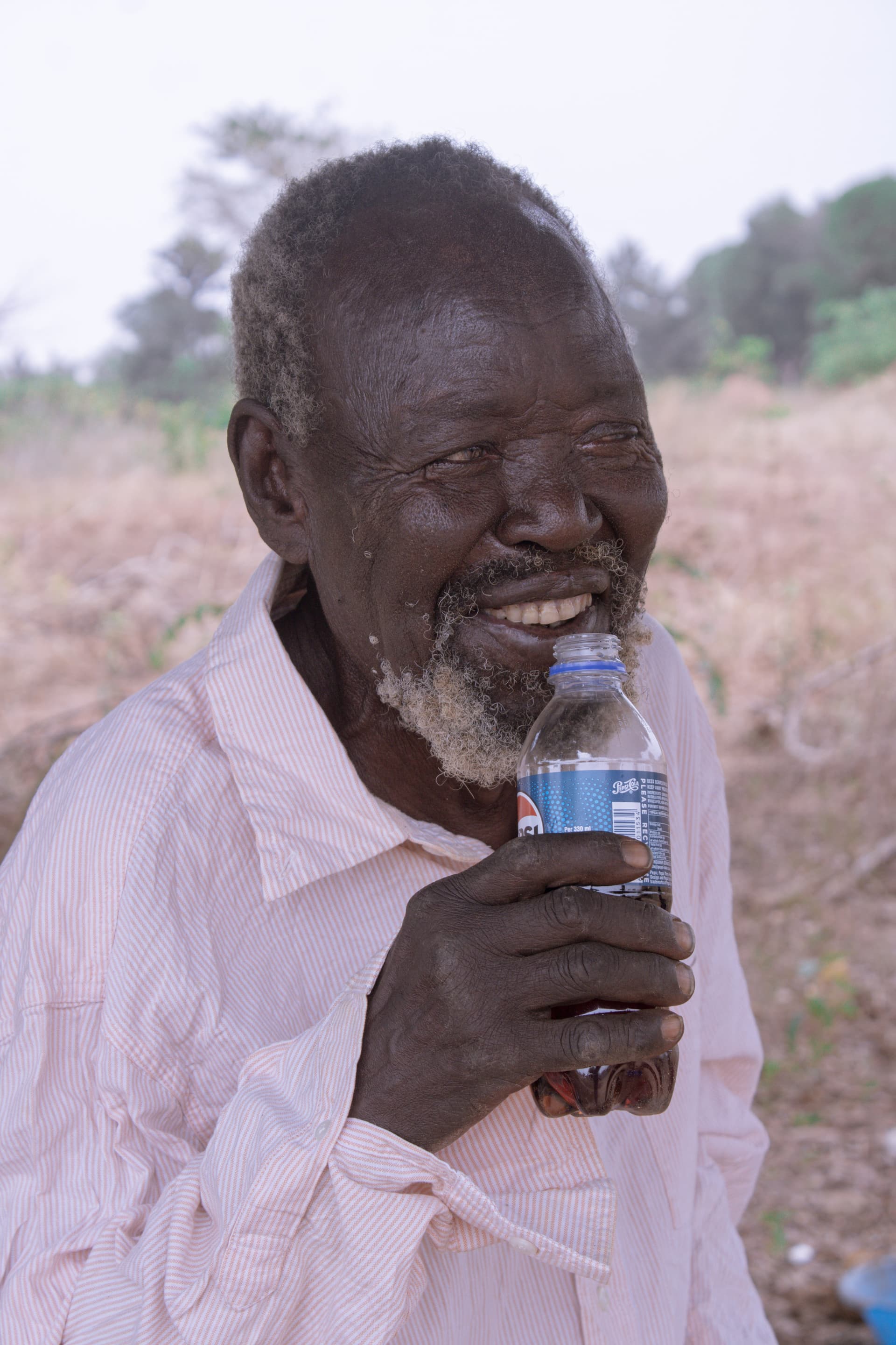 John, a 90-year-old man, smiles while drinking from a plastic water bottle outdoors.