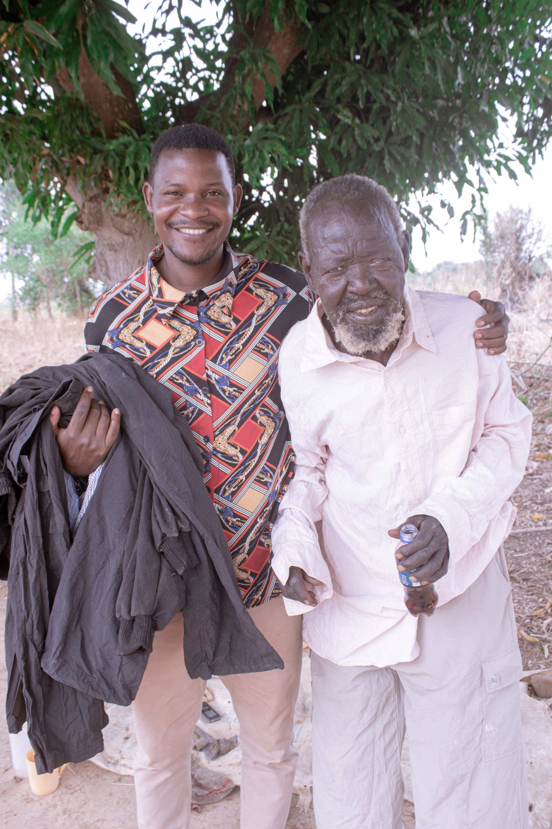 A JFM worker presents new clothing to John, the elderly man, both smiling outdoors.