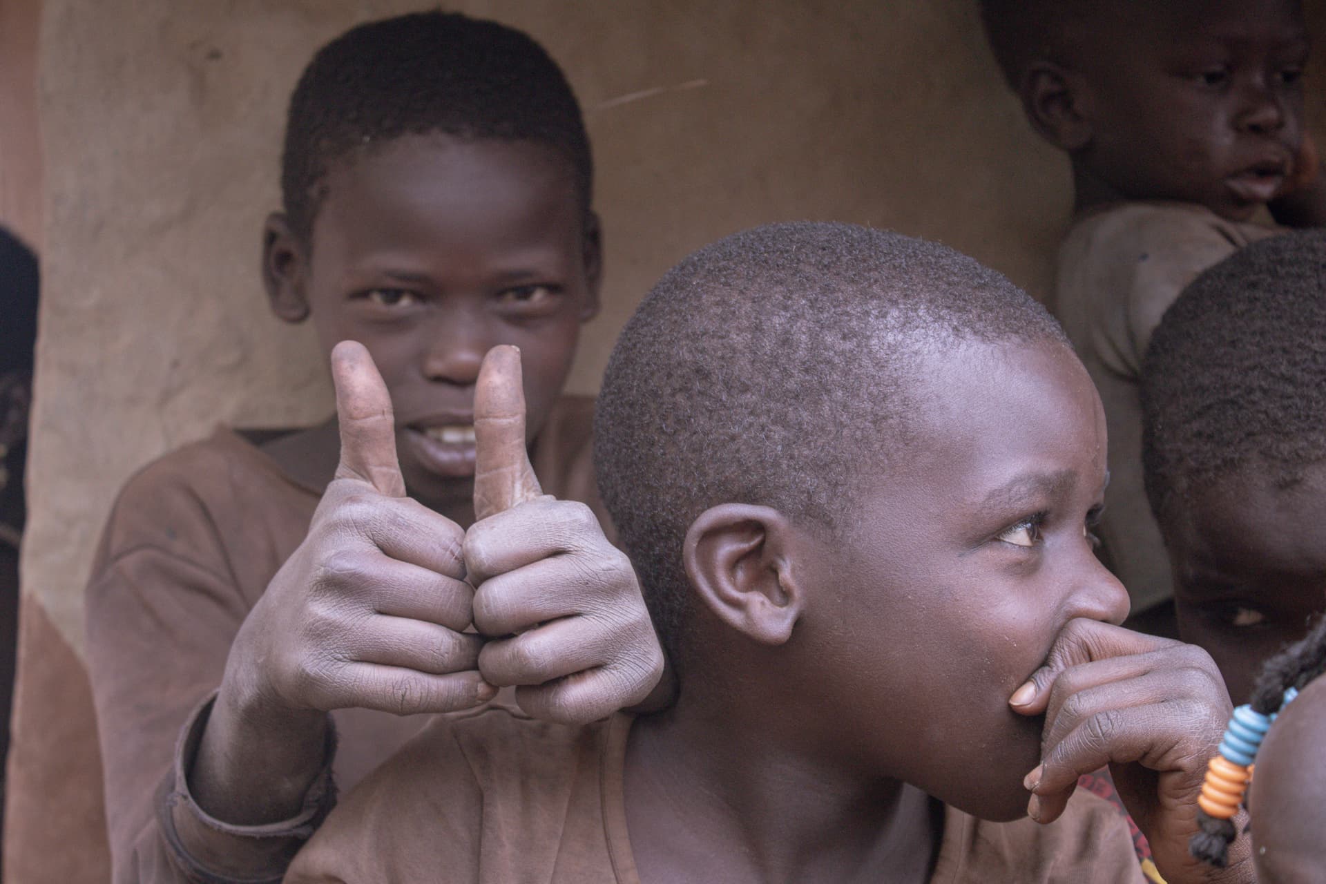 Two boys stand close together outdoors, one giving a thumbs-up and smiling at the camera.
