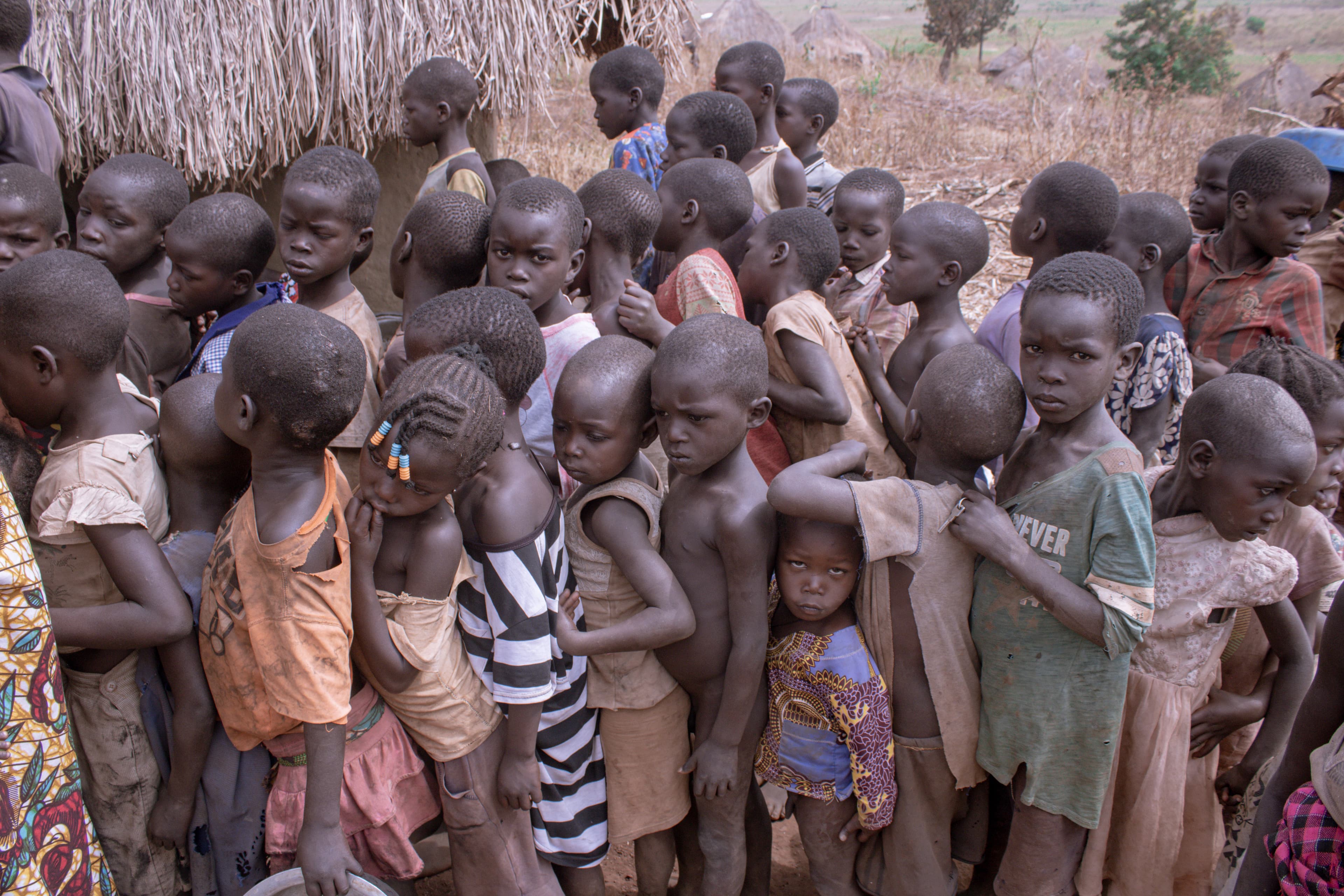 A large group of children stands together outdoors near a thatched structure.
