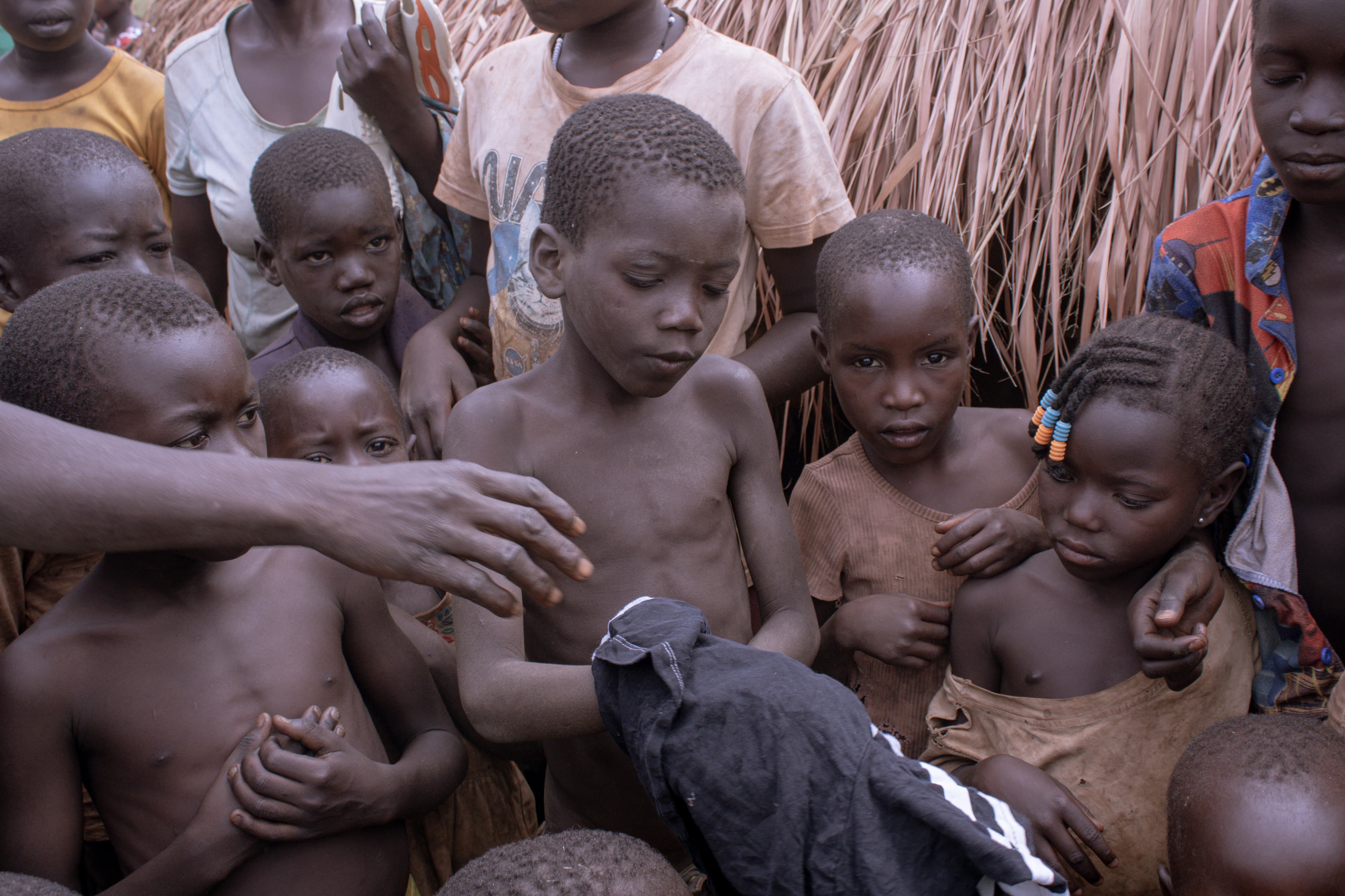 An adult reaches toward a group of children, one wrapped in a dark cloth, near a thatched hut.