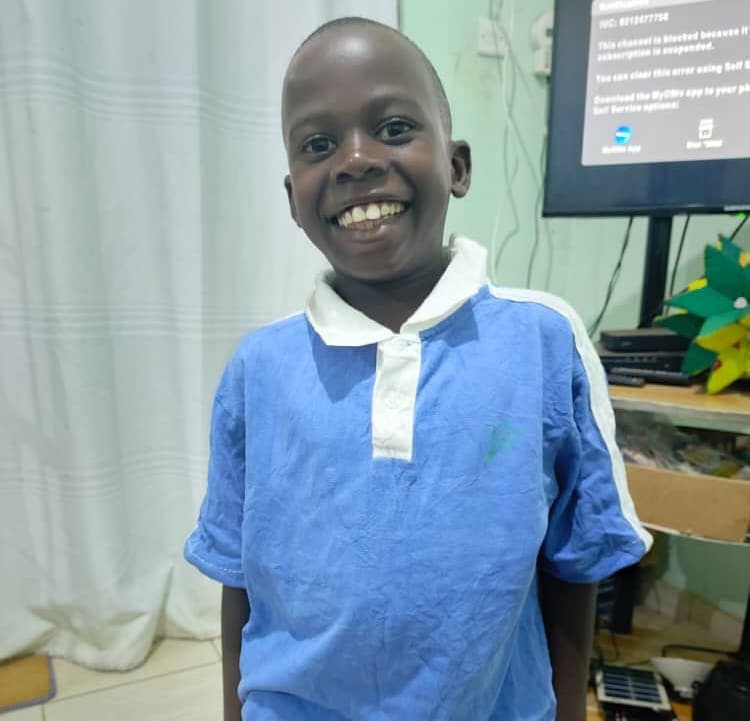 Boy in a blue polo shirt smiling indoors near a screen or monitor.