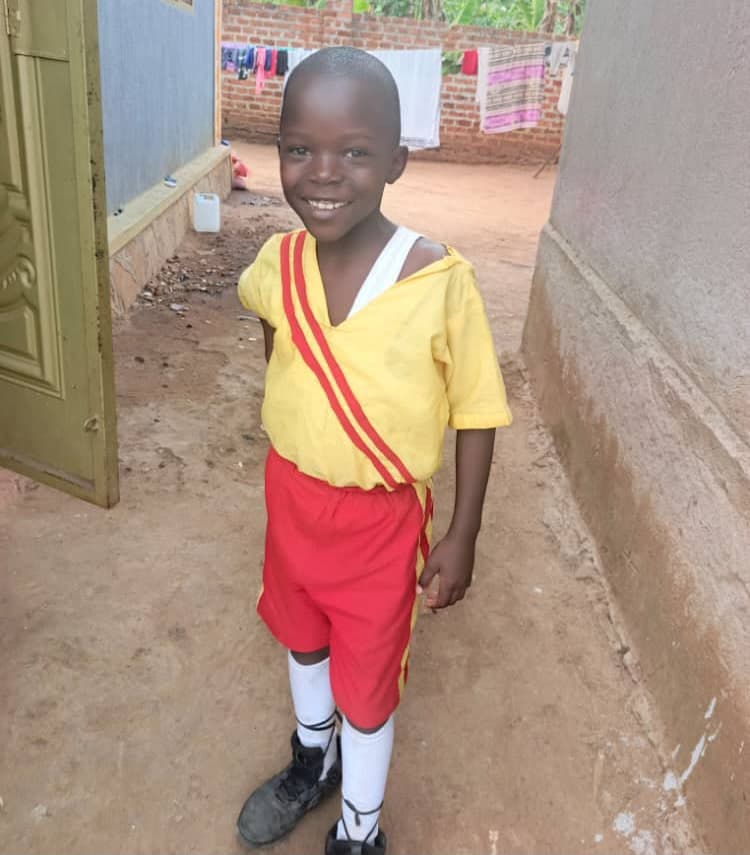 Young boy in a yellow shirt and red trousers standing indoors, part of a school uniform.