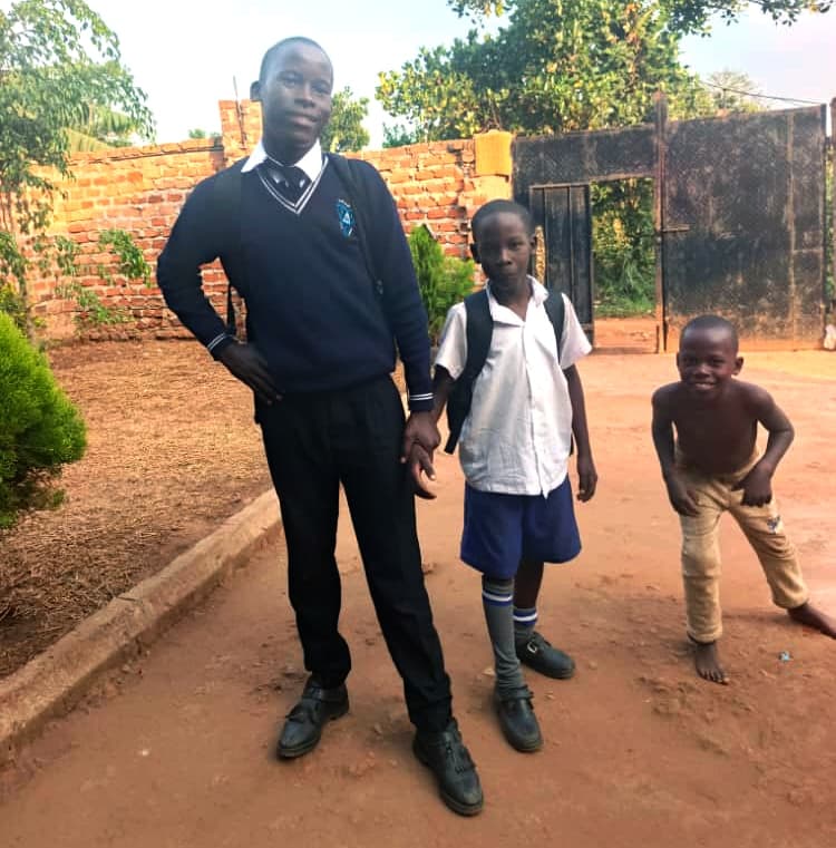 Older boy in a school uniform standing with younger children outside a brick building.