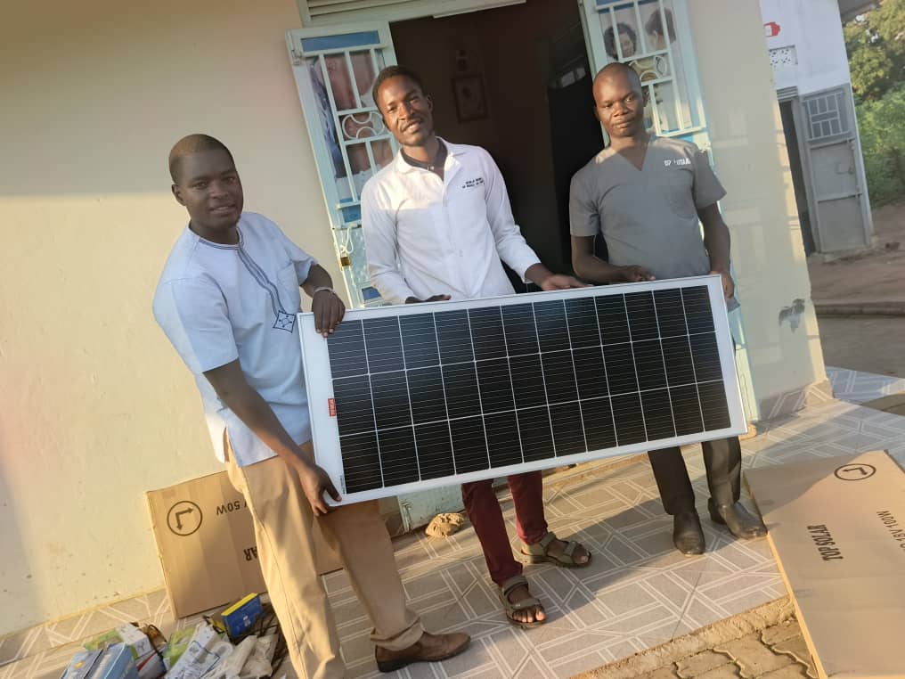 Three men stand outside Miracle Medical Center holding a solar panel, with boxes on the ground.