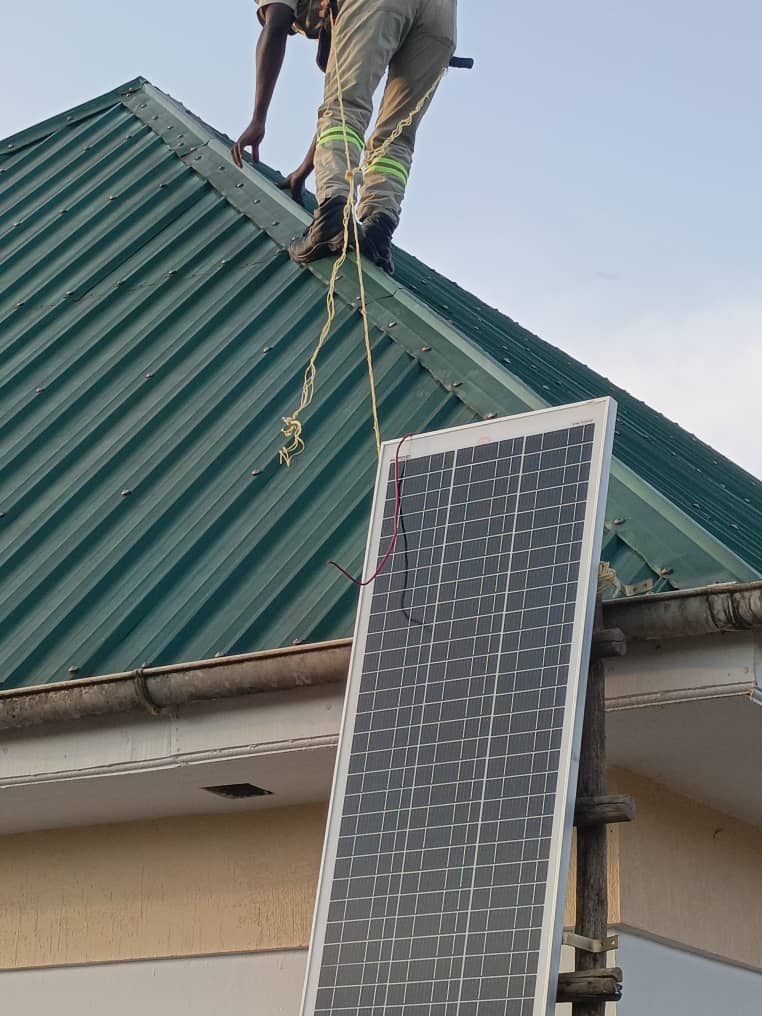 A worker on a rooftop lowers a solar panel by rope to someone below.