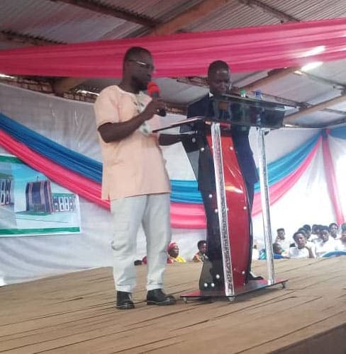 A man speaks at a podium under a pink and blue event tent, with a second man standing beside him.