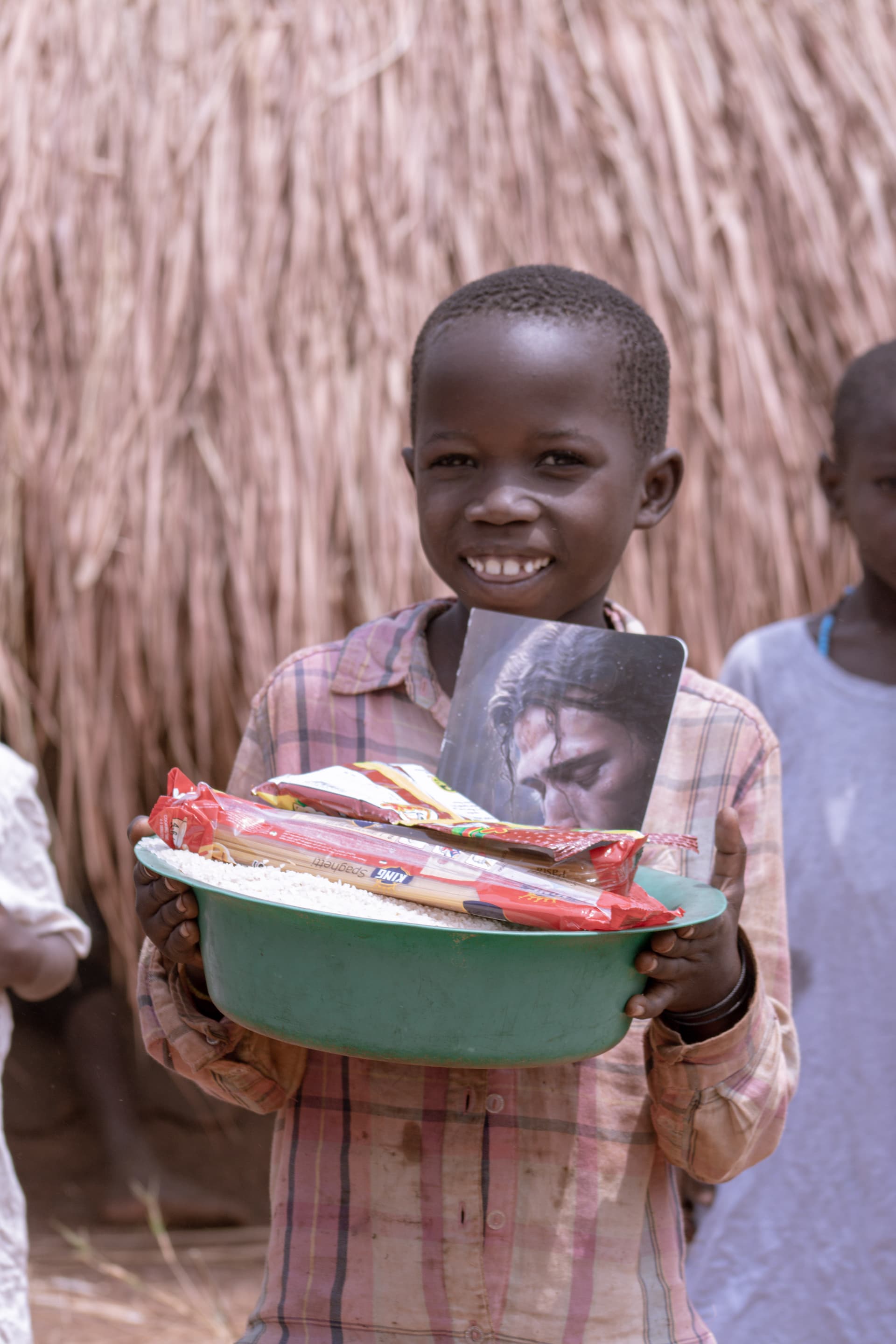 A smiling boy holding a green and white basin filled with food packets, standing by a thatched hut.
