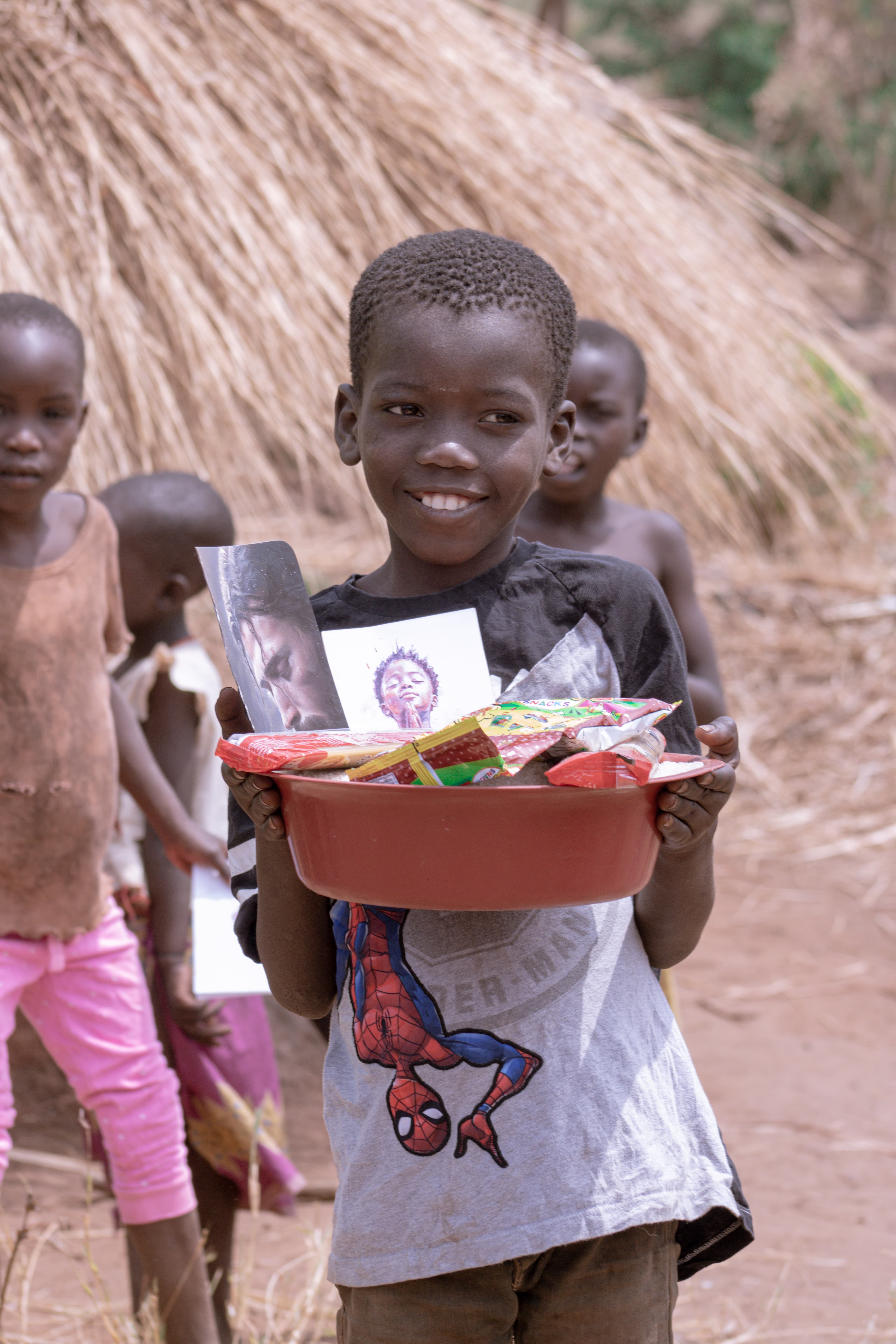 Boy holding a red basin of food and printed materials, with other children nearby.