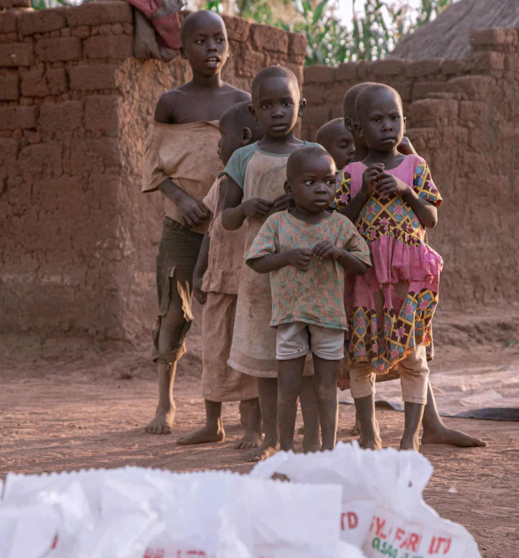 Group of children stand near a mud building beside stacked bags of food supplies.