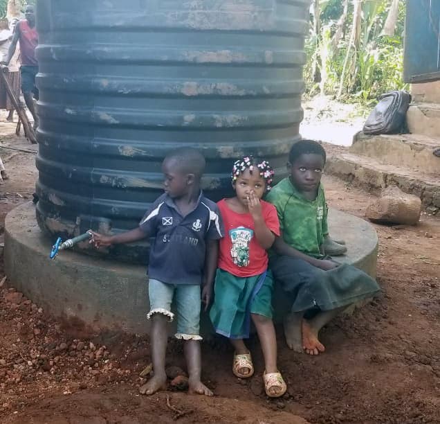 Children in red school uniforms stand beside a large black water tank outside a brick building.