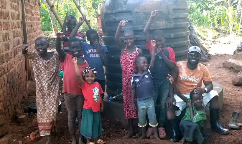 Group of children and adults stand near a large black water tank among trees.