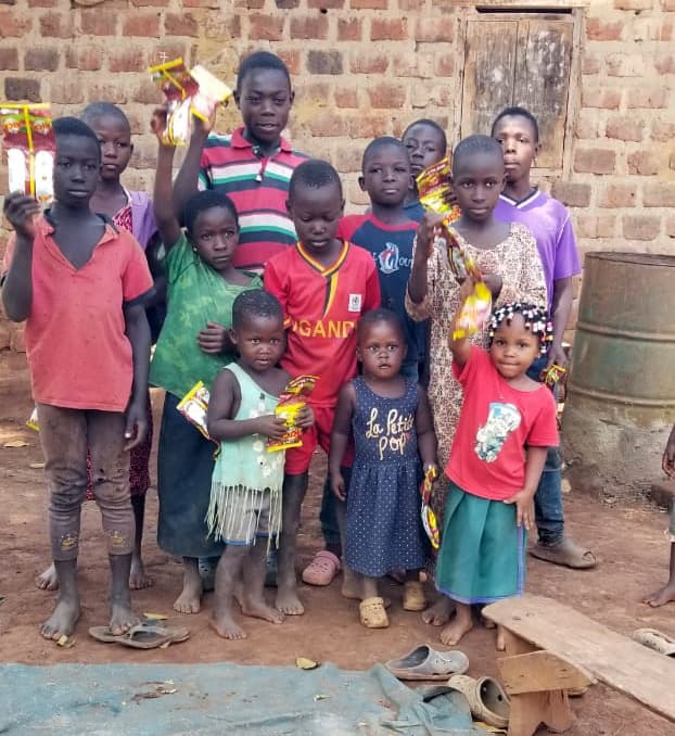 Children in red and green uniforms stand near a brick building and water barrel.