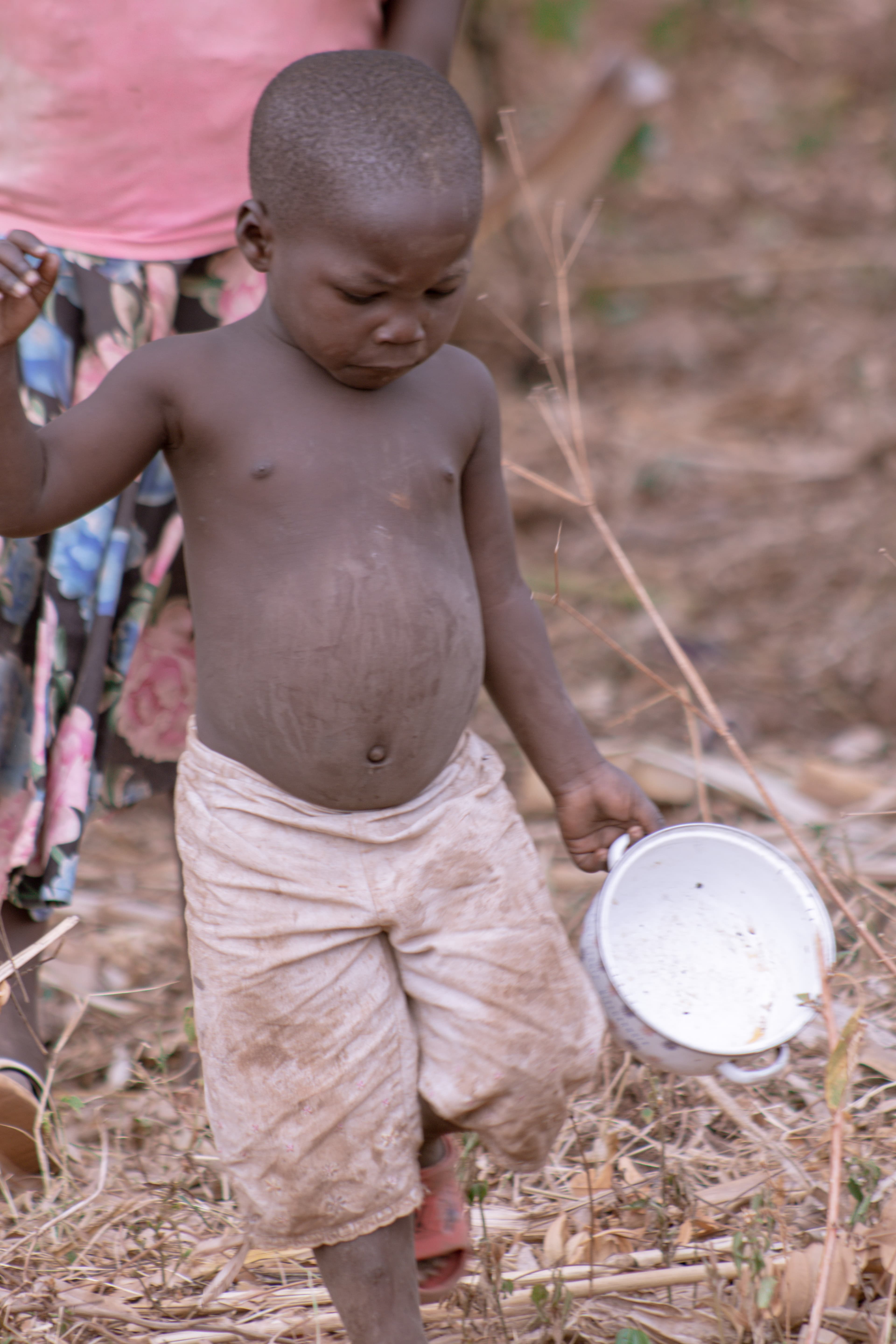 A young shirtless boy standing with a white cup or bowl in a dry field.