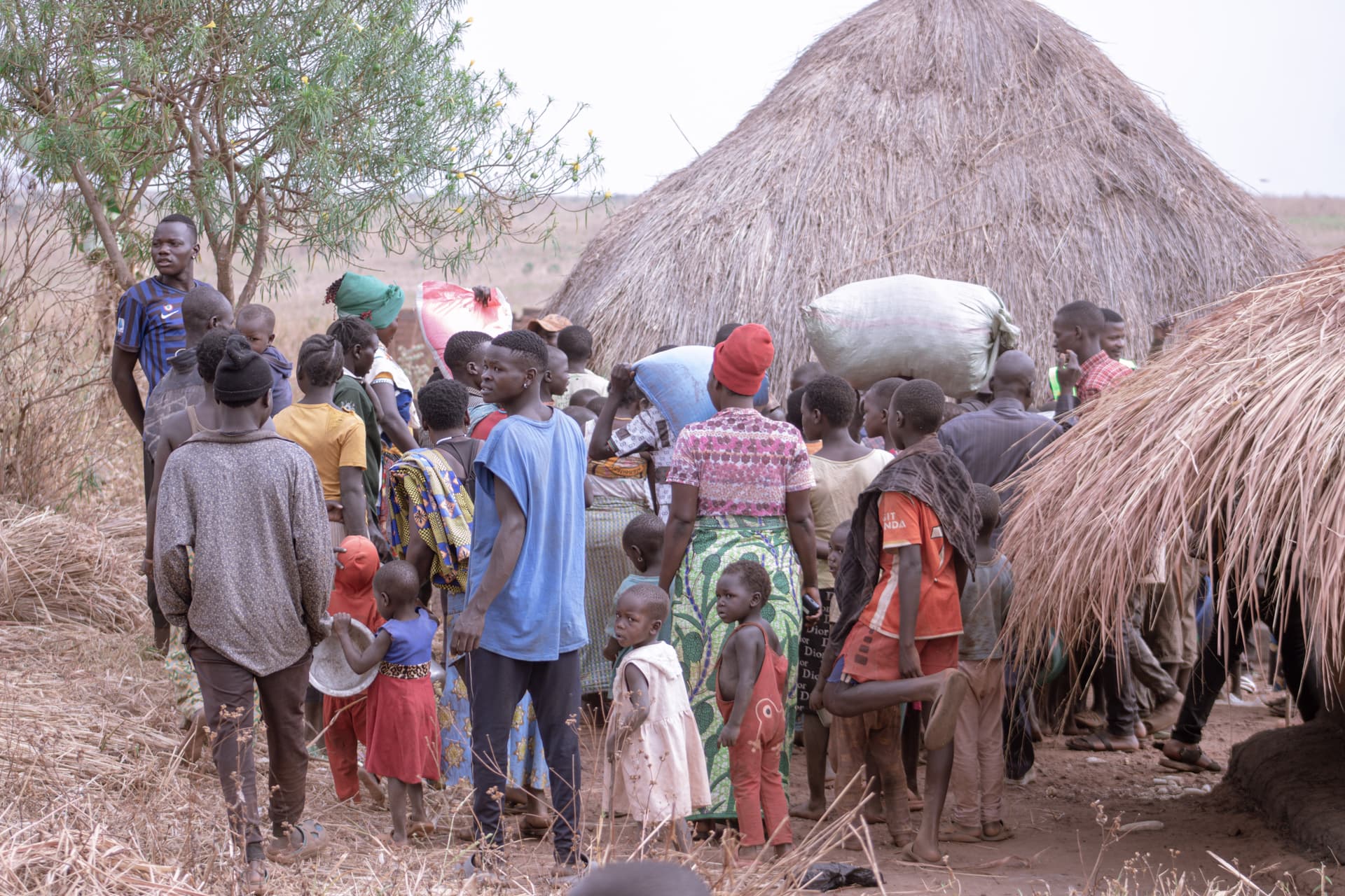 A group of people walking single file past thatched huts carrying belongings.