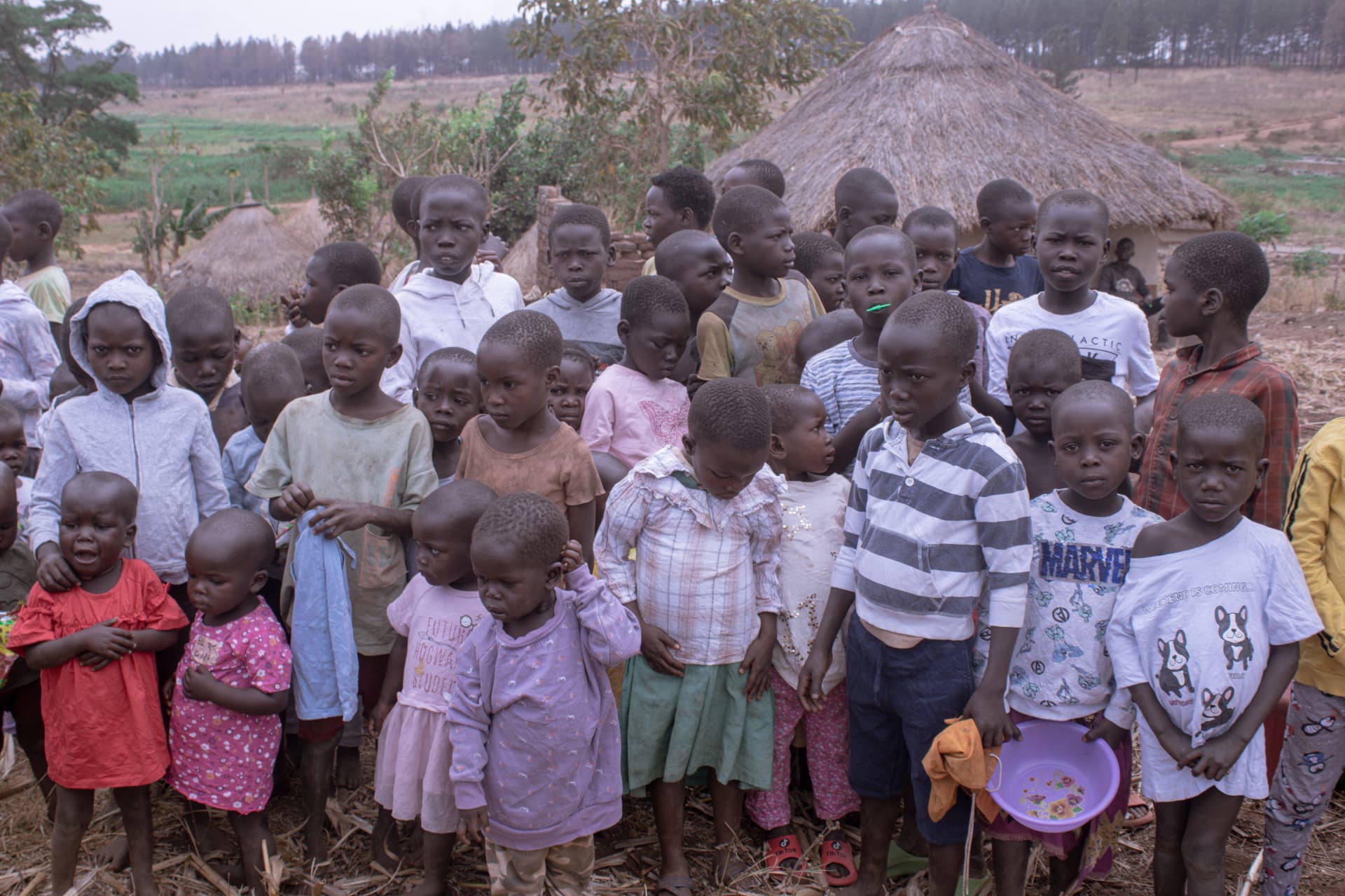 A large group of children and women gathered outdoors near thatched huts.