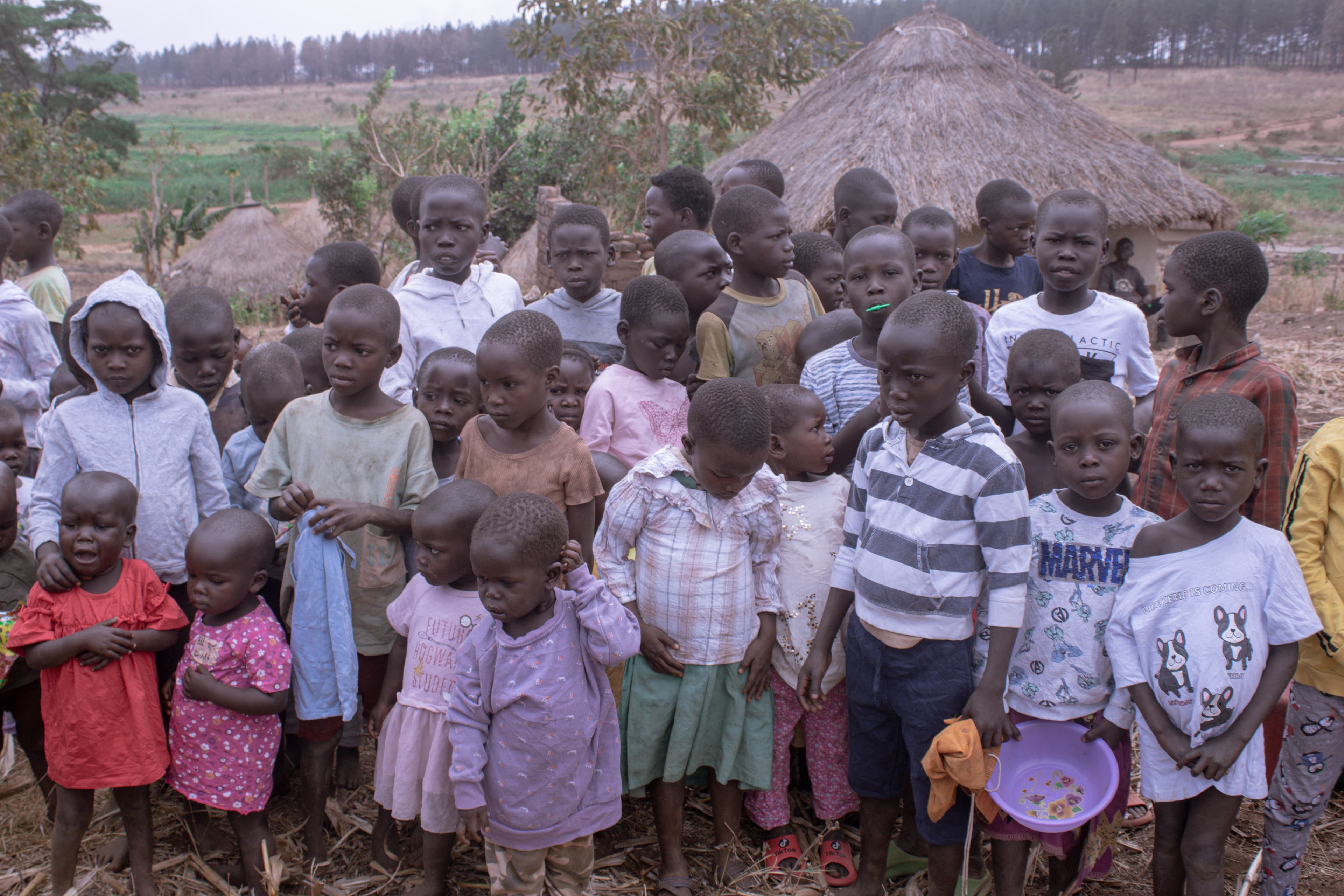 A crowd of children gathered in front of a thatched-roof home during a village visit.
