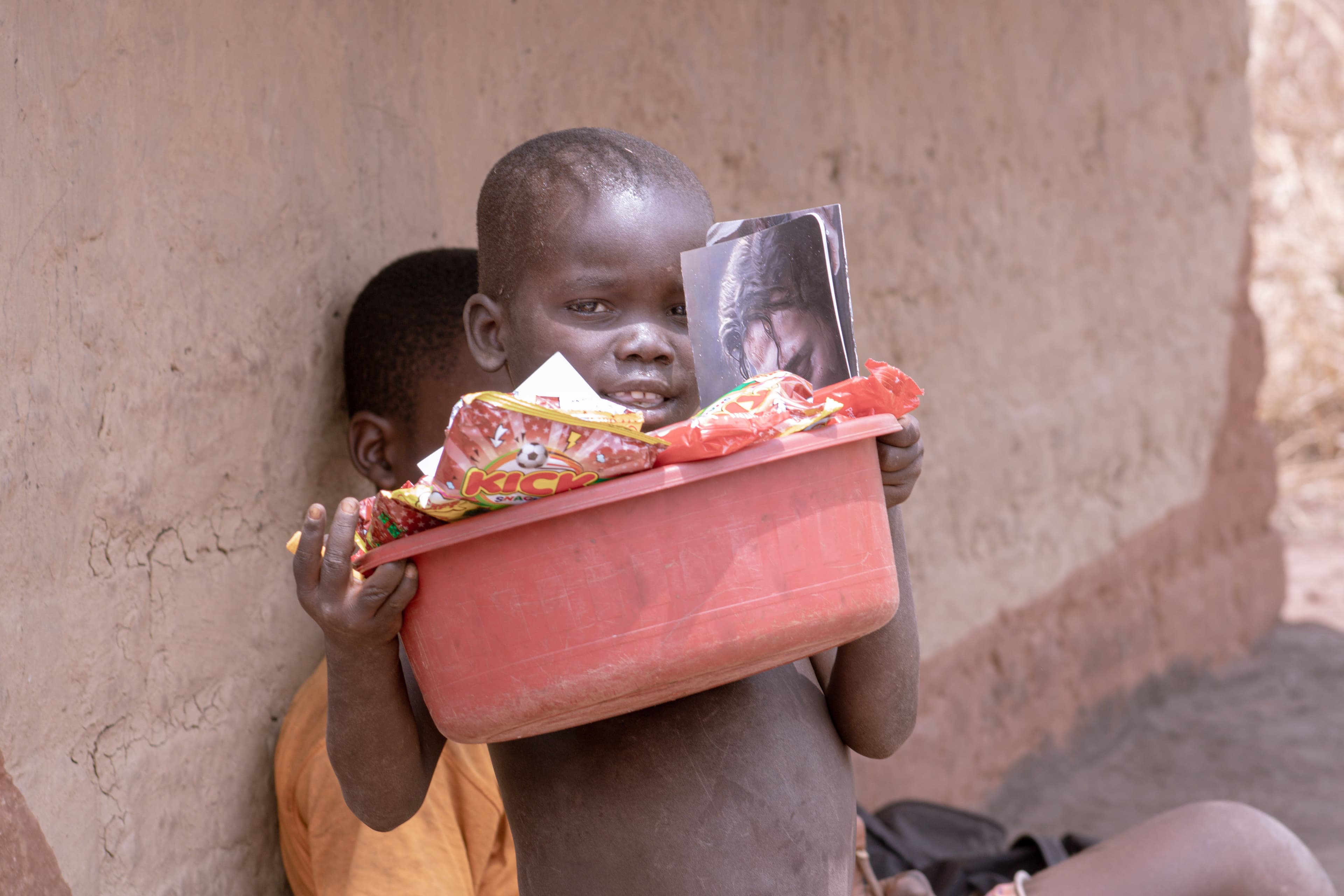 A young child holding a red basin of food gift items against a mud wall.