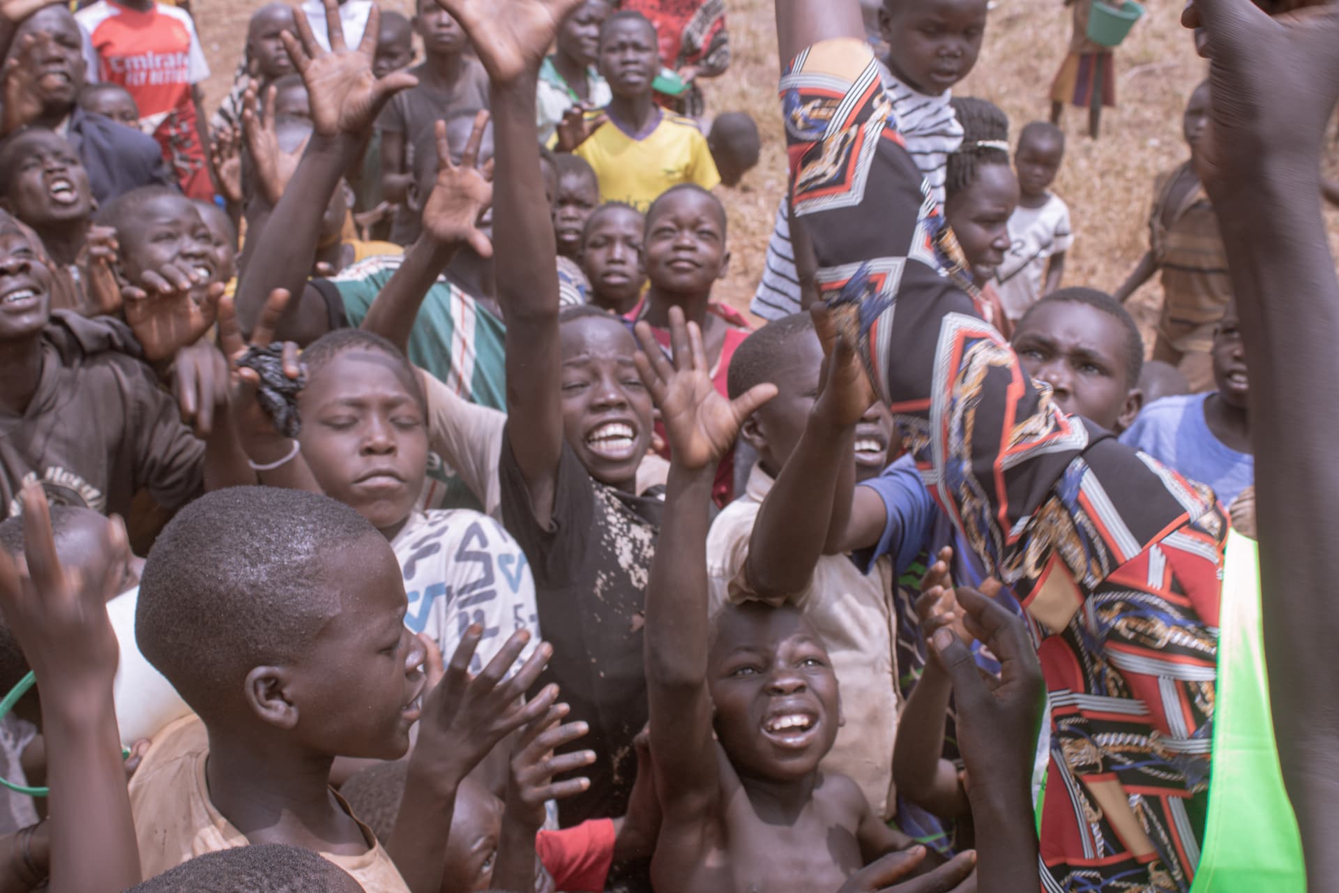 A crowd of children raising their hands and cheering, some wearing colorful traditional cloth.