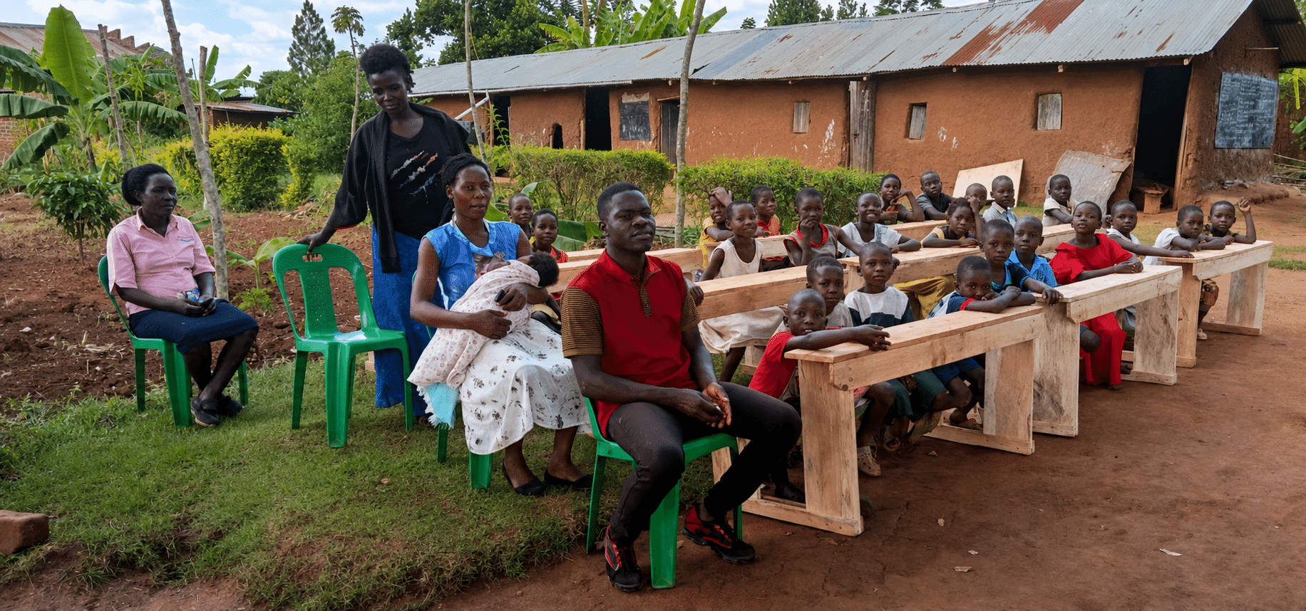 Students and a teacher sit at wooden desks set up outdoors beside a school building.