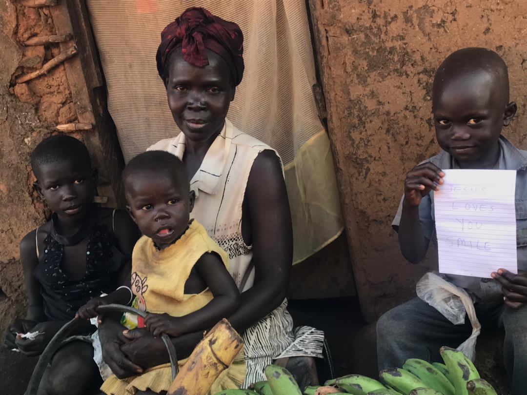 Mother and children standing outdoors, an older child holding a handwritten note beside a banana bunch.