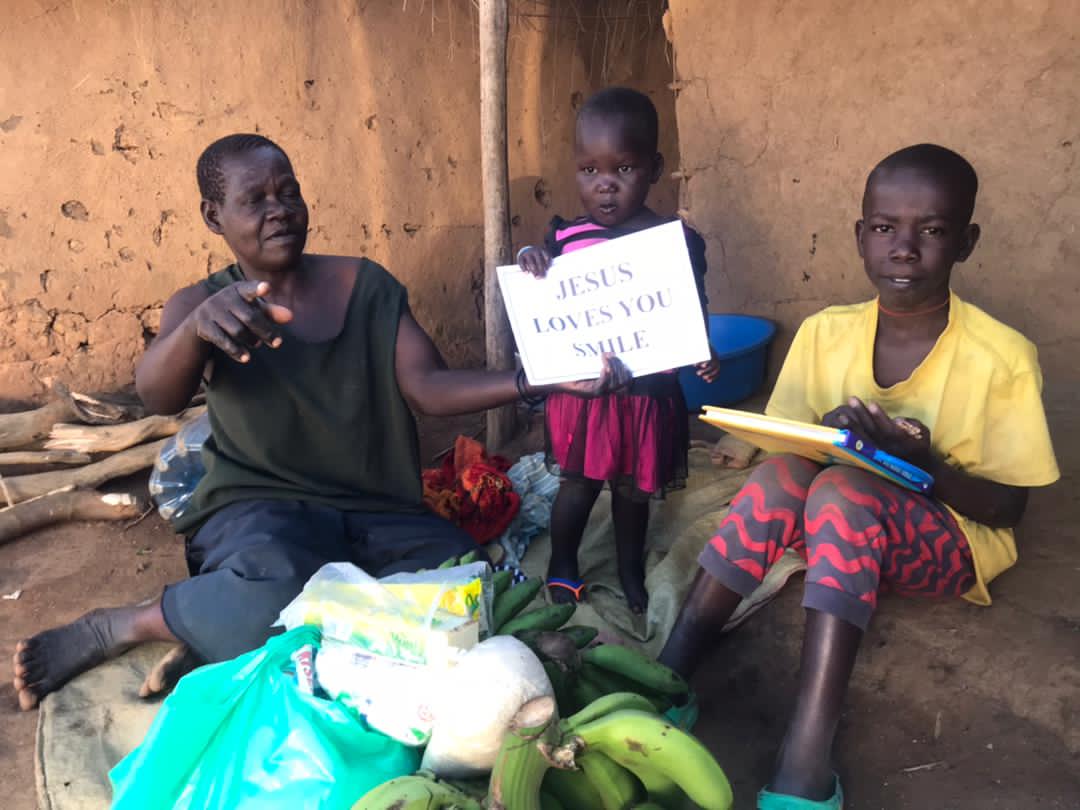 Akello sits against a mud-brick wall holding a Jesus Fed Me gospel tract sign beside a girl in a yellow shirt.