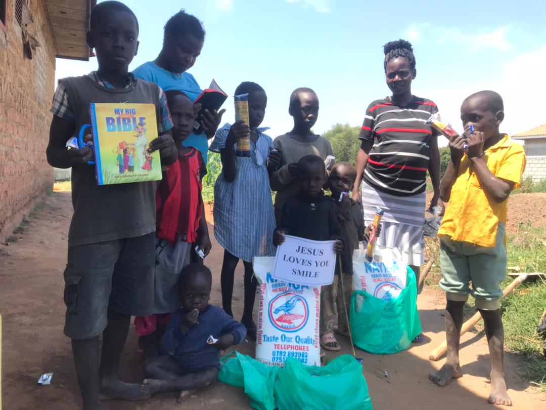 Teenage mothers and children standing outdoors holding Bibles and a green food relief bag.