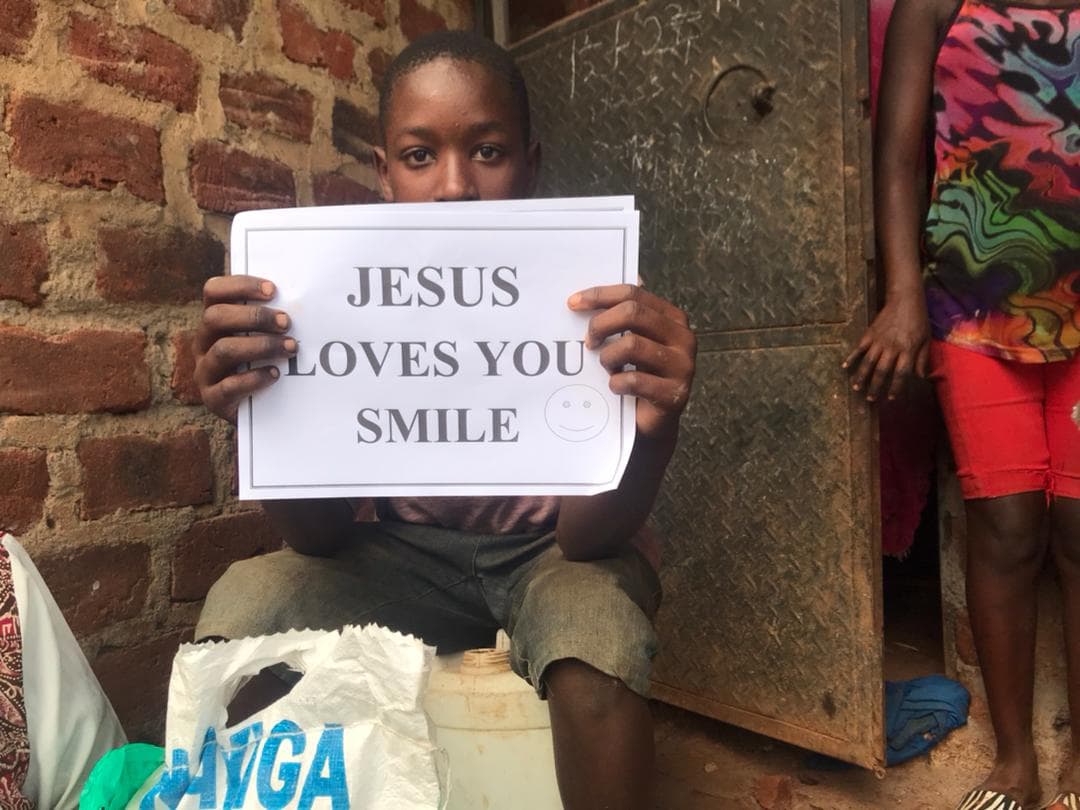 Child holding a 'Jesus Loves You Smile' sign while seated near a brick wall.