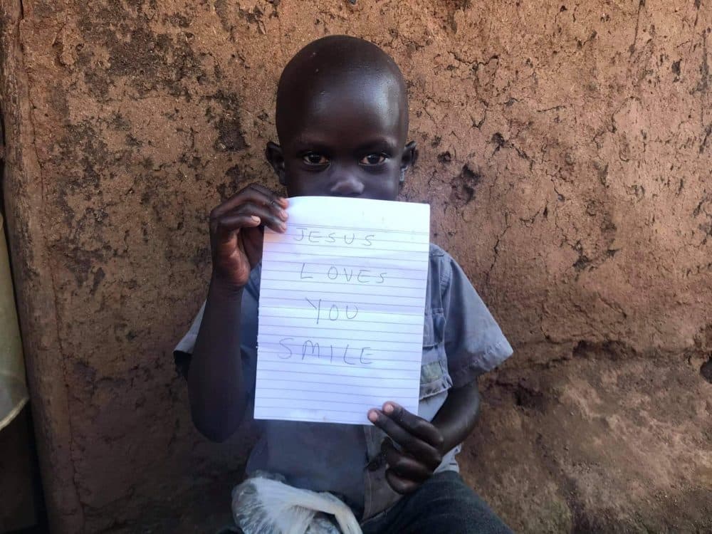 A young child sits against a mud wall holding up a handwritten Jesus Fed Me note, partly covering their face.