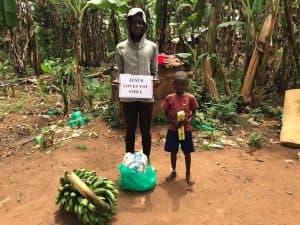 Backlit photo of a woman and child standing outdoors holding a 'Jesus Loves You Smile' sign beside a banana bunch.