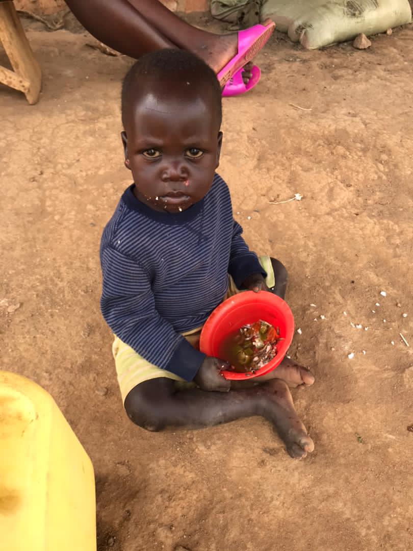 Toddler sitting outdoors on the ground holding a red bowl of food and looking at the camera.