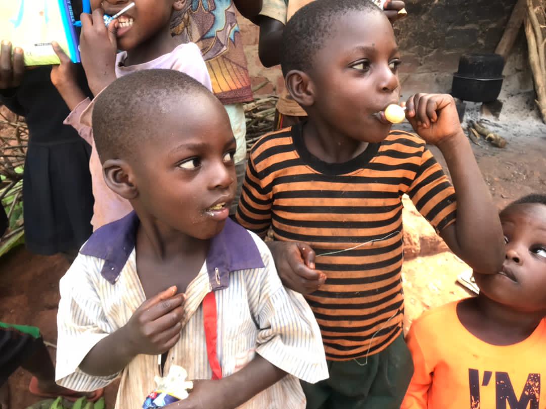 Two boys stand outdoors, one holding a chewing stick or toothbrush in his mouth.