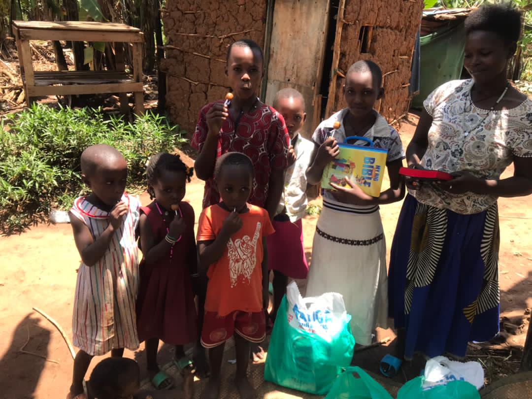 Mrs. Olivia and her children stand outdoors near a mud-brick wall holding relief items.