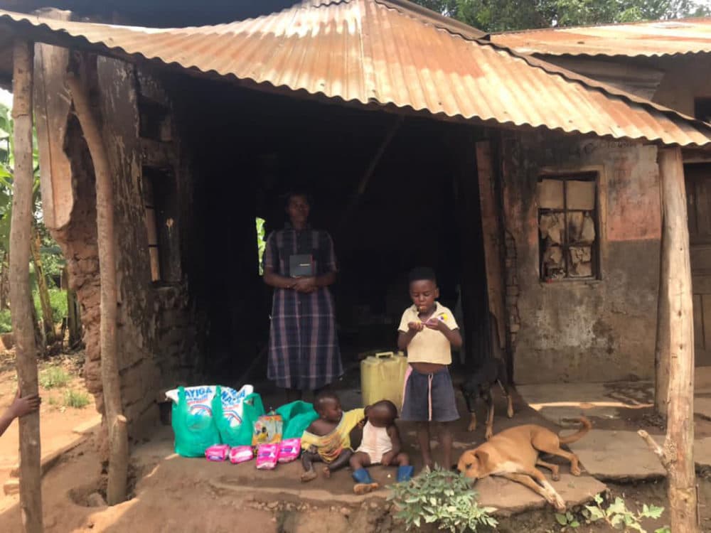 A man stands in the doorway of a small thatched-roof mud house with a woman and child, green gift bags on the ground.