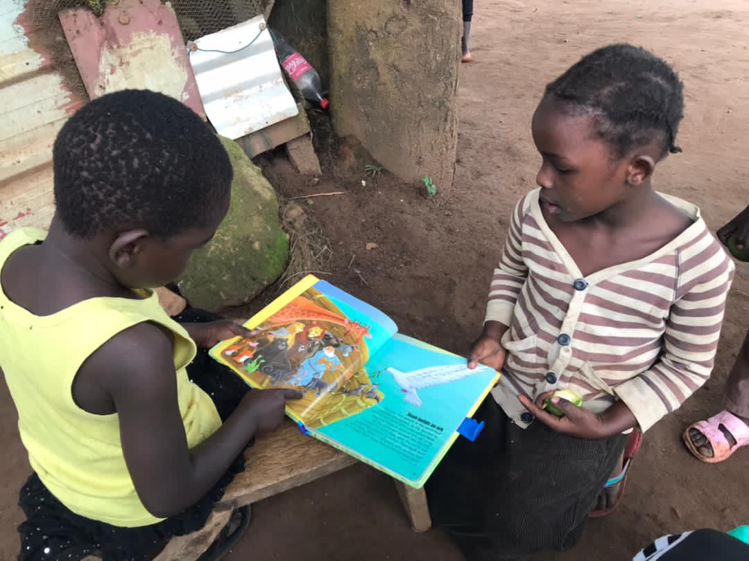 Two of Mrs. Monica's children sit together looking at an open picture Bible book.