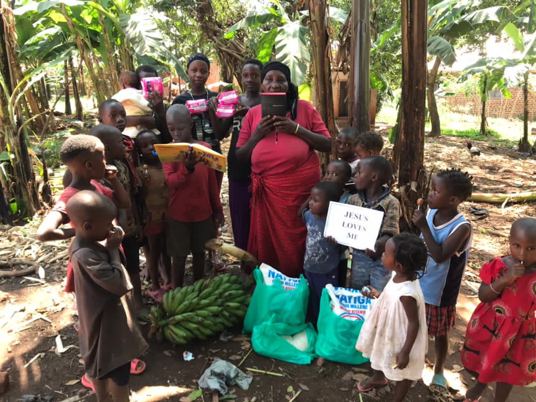 Grandmother Christine, in a red dress, stands with a large group of grandchildren under banana trees holding gift bags.