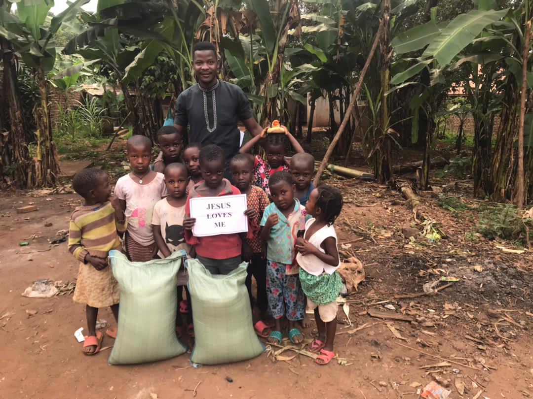 A man and several children stand outside a brick house beside large sacks of rice, one child holding a paper sign.
