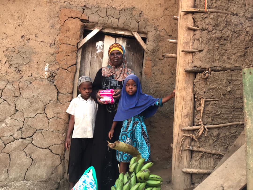 Two women and a child stand in the doorway of a mud-brick house with a bunch of bananas in the foreground.