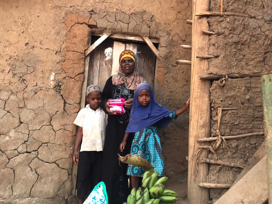 Mrs. Aidah, in a hijab, stands with her two children in the doorway of their mud-brick home, one child holding a gift bag of bananas.